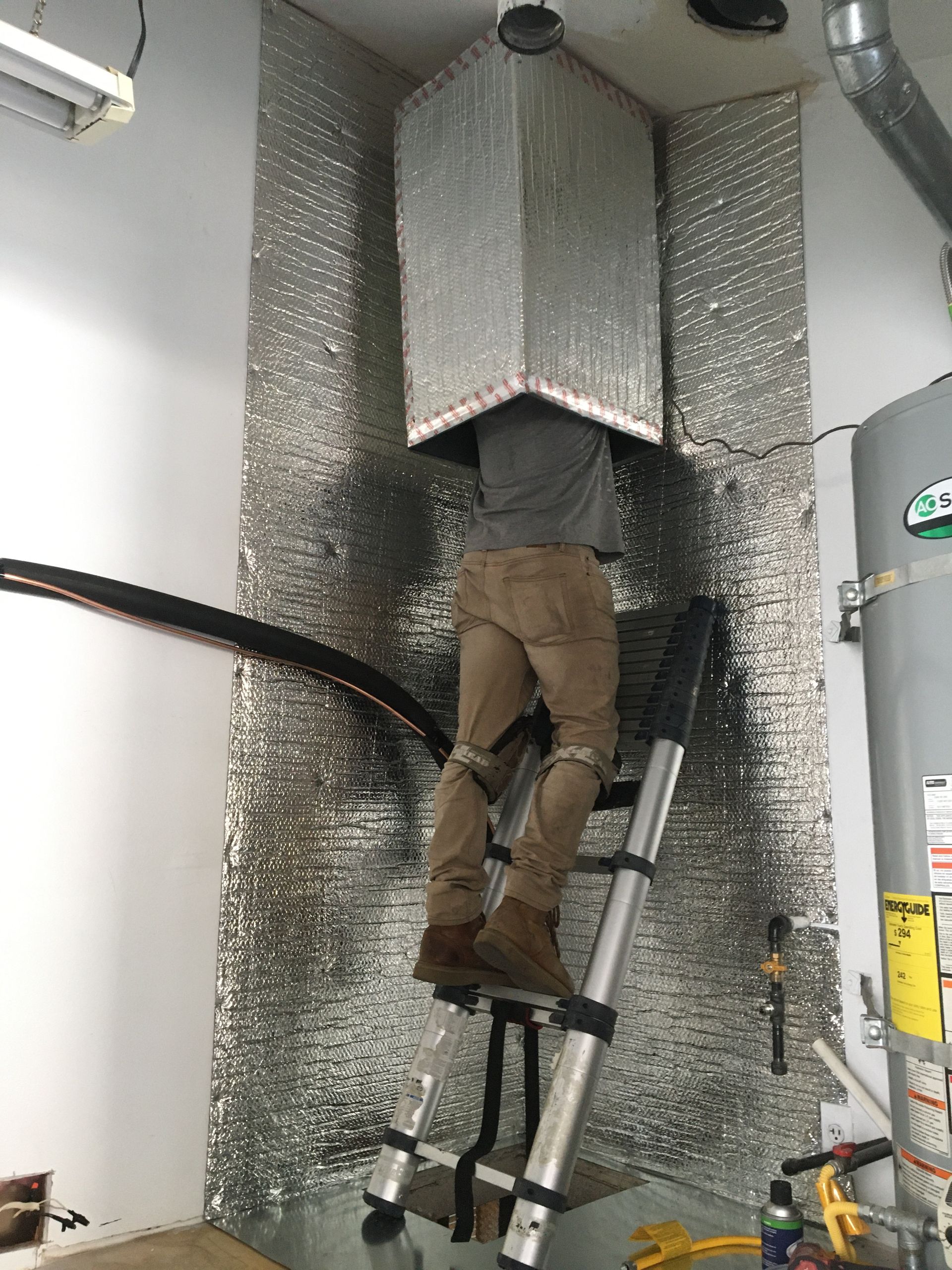 Person on a ladder installing insulation in a utility room with silver foil-covered ducts and pipes.