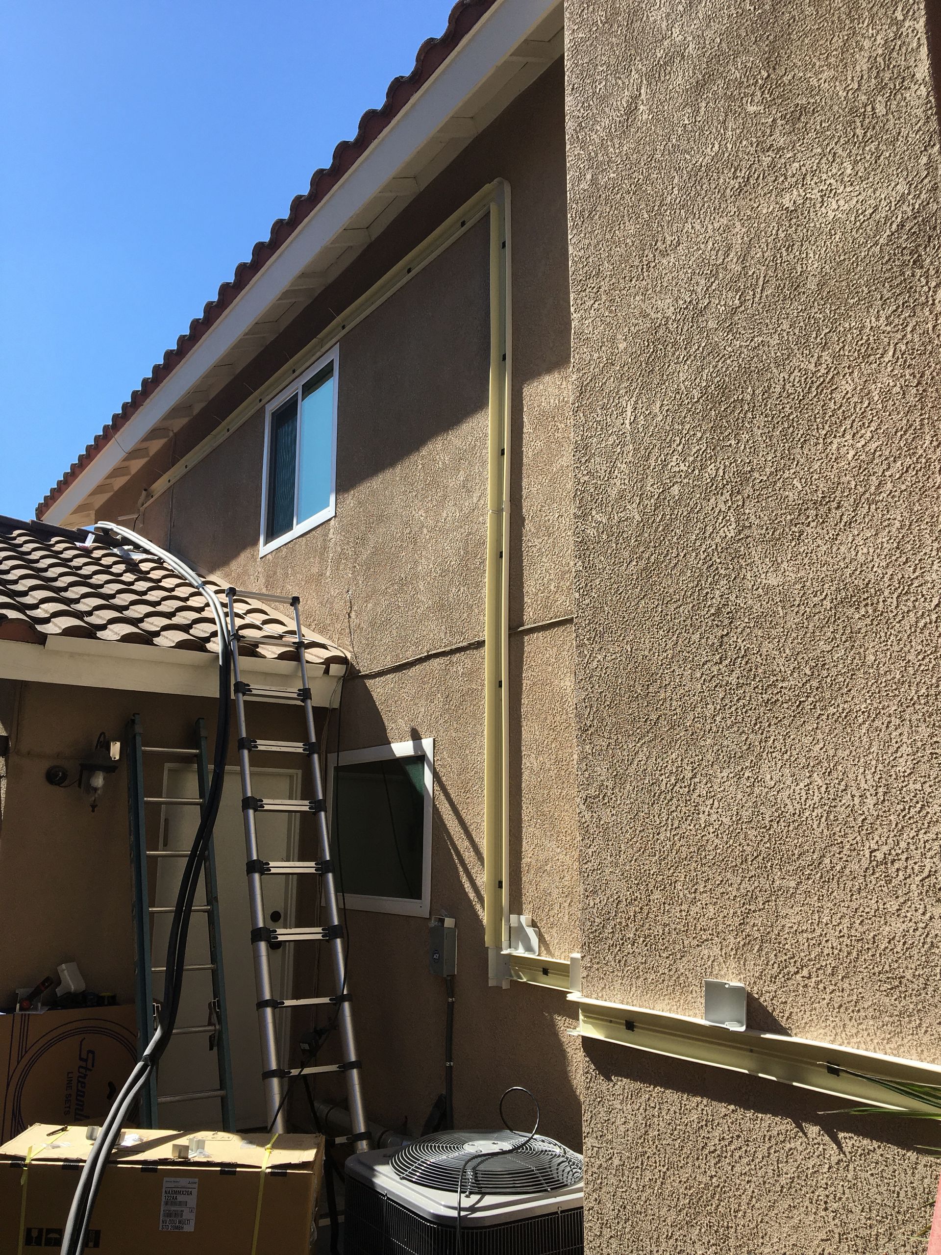 Backyard view of a stucco house wall, ladder, roof edge, and a small window under a blue sky