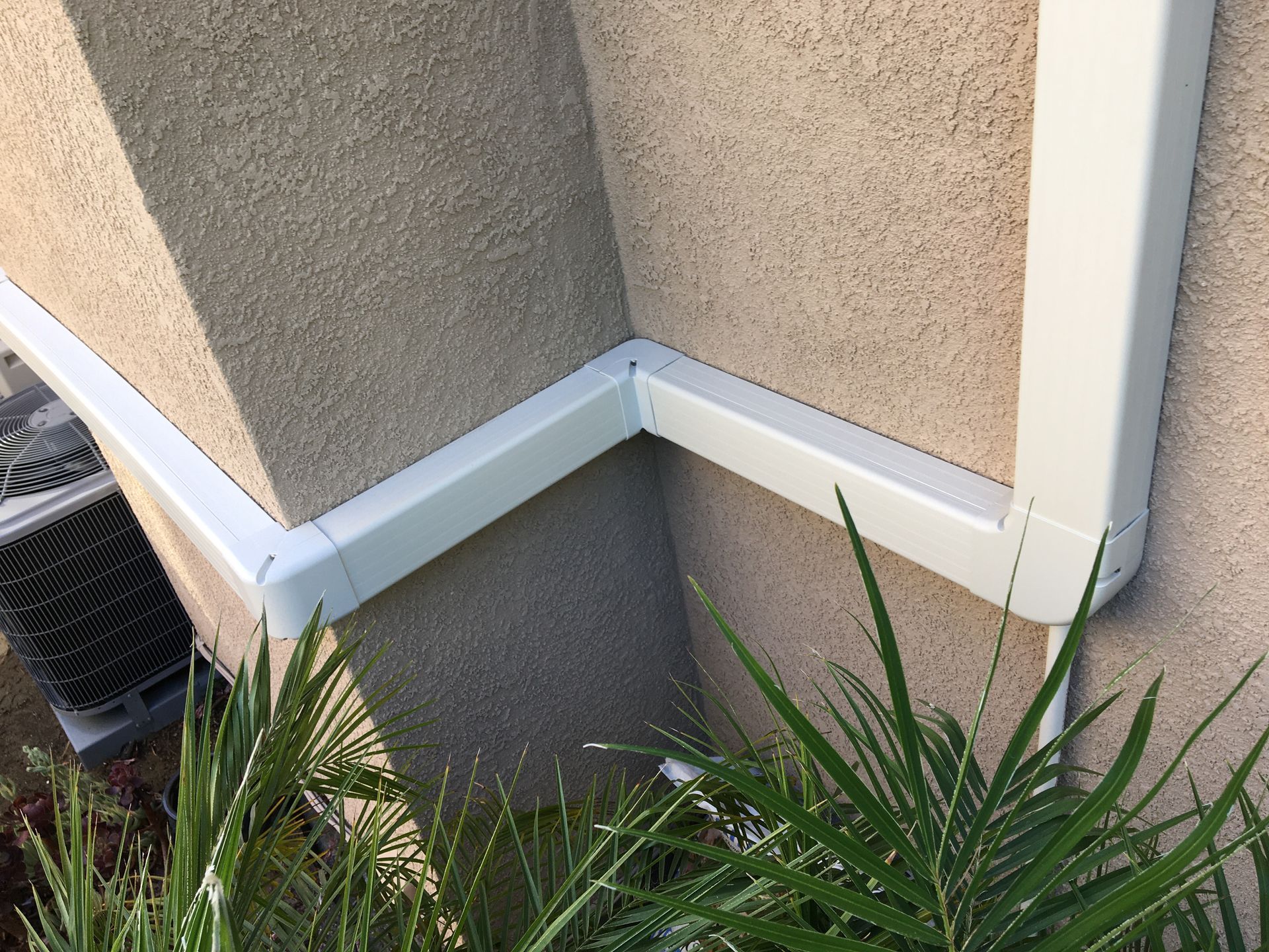 White downspout on beige stucco house corner above grass and plants
