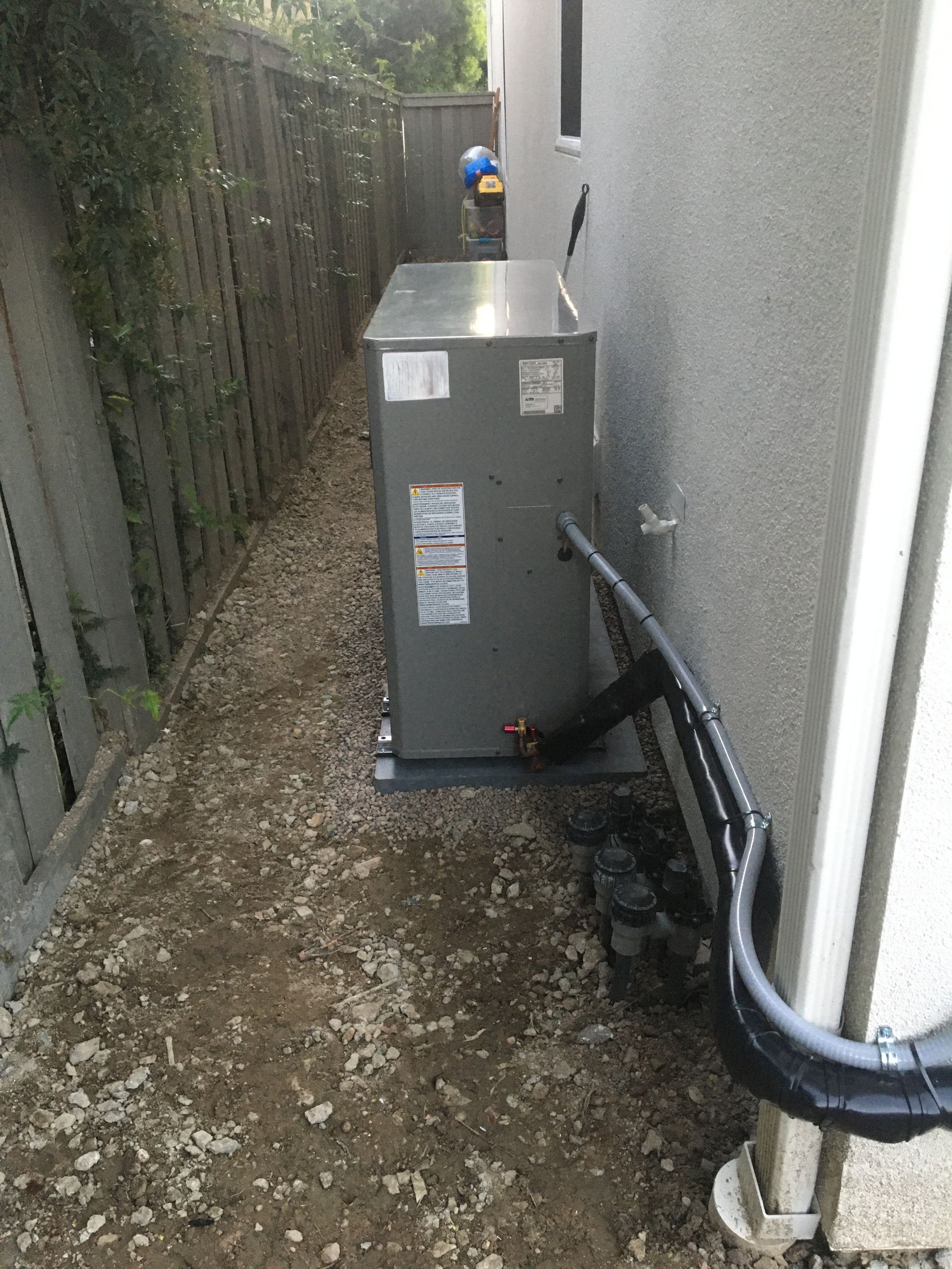 Narrow outdoor utility alley with a gray HVAC unit beside a white wall and a fence-lined gravel path