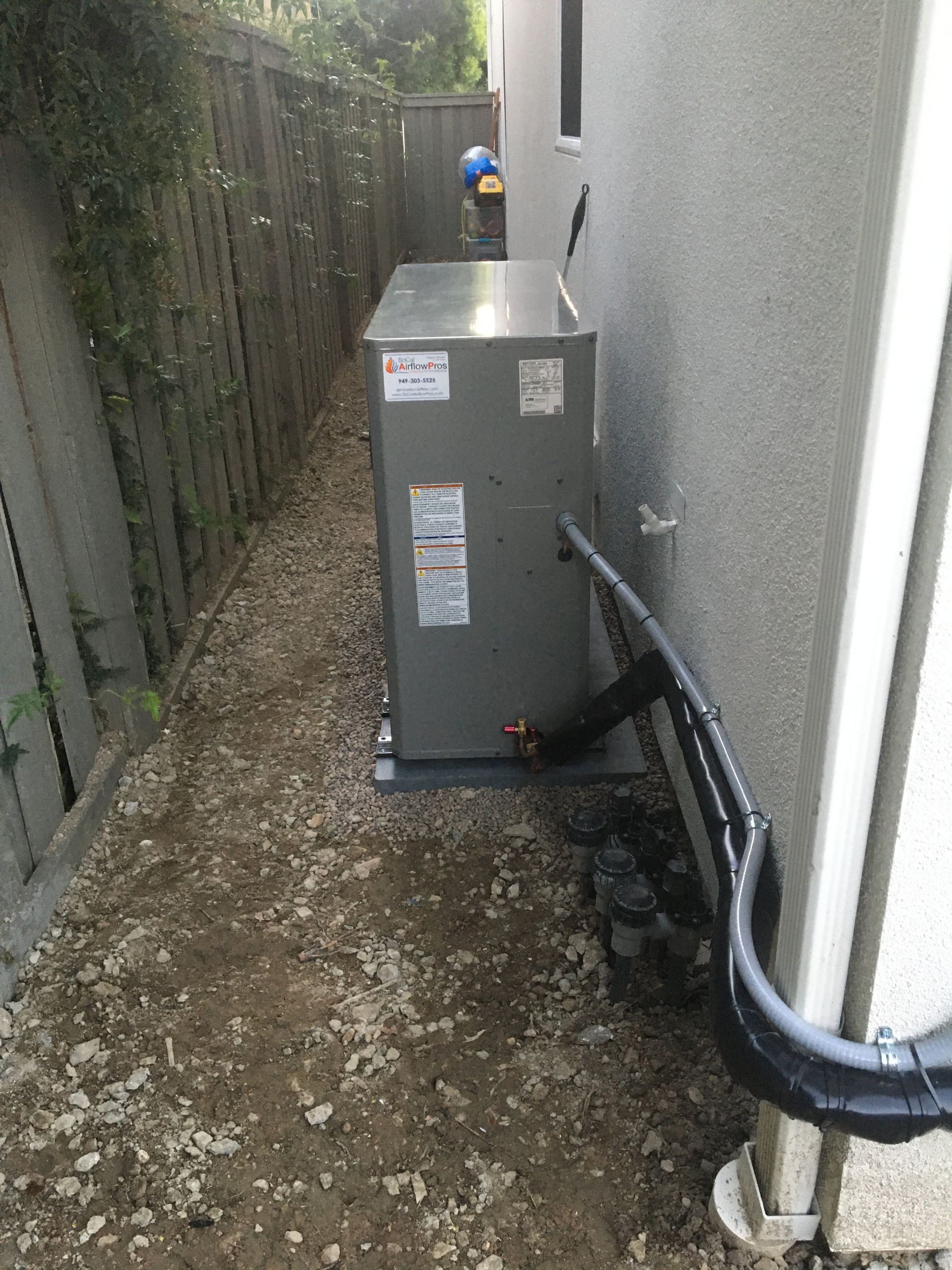 Narrow side yard with a gray utility unit, pipes, gravel ground, and a person in the distance.