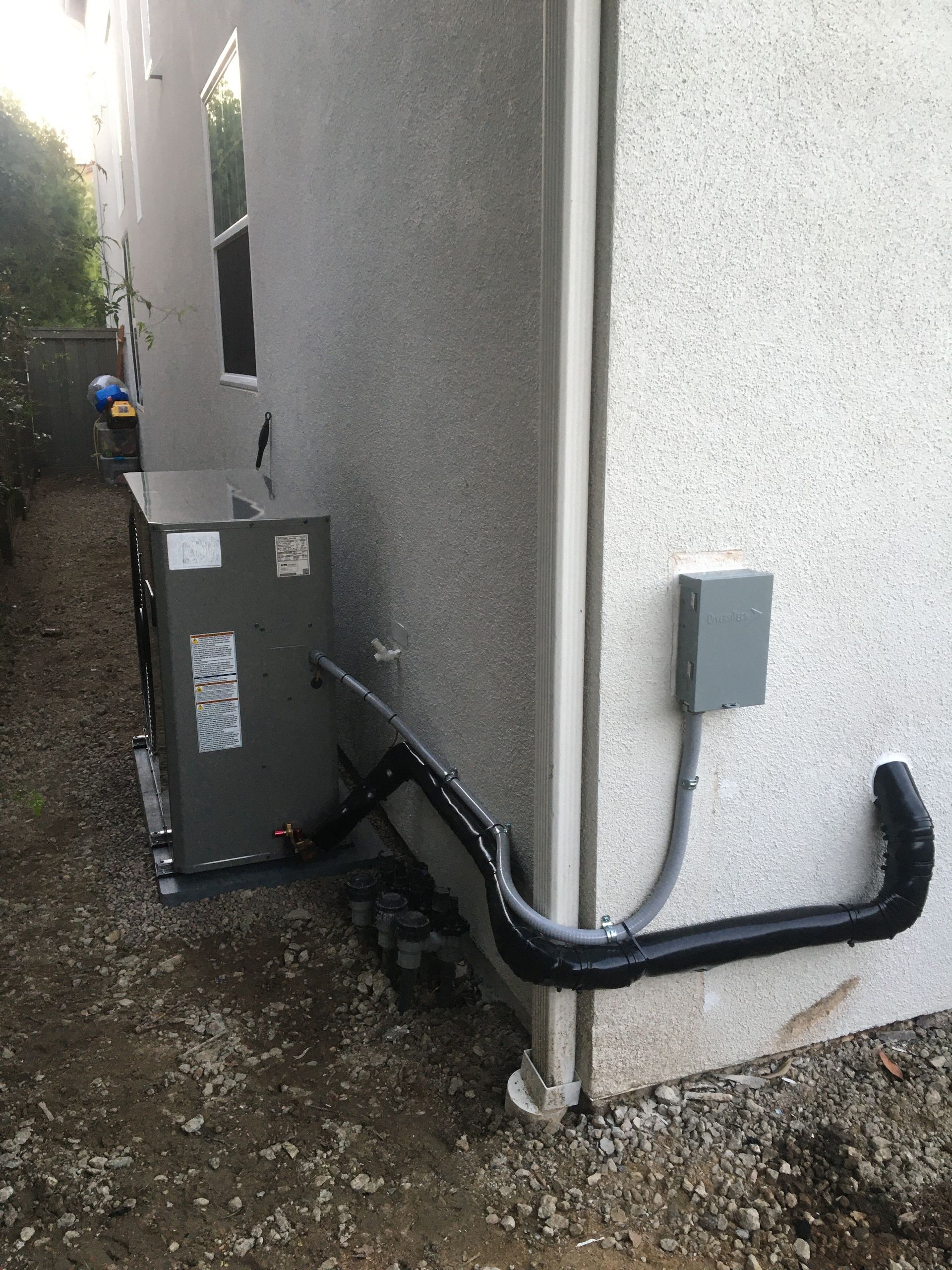 Narrow side yard with utility boxes, exposed pipes, and a black conduit along a stucco wall.