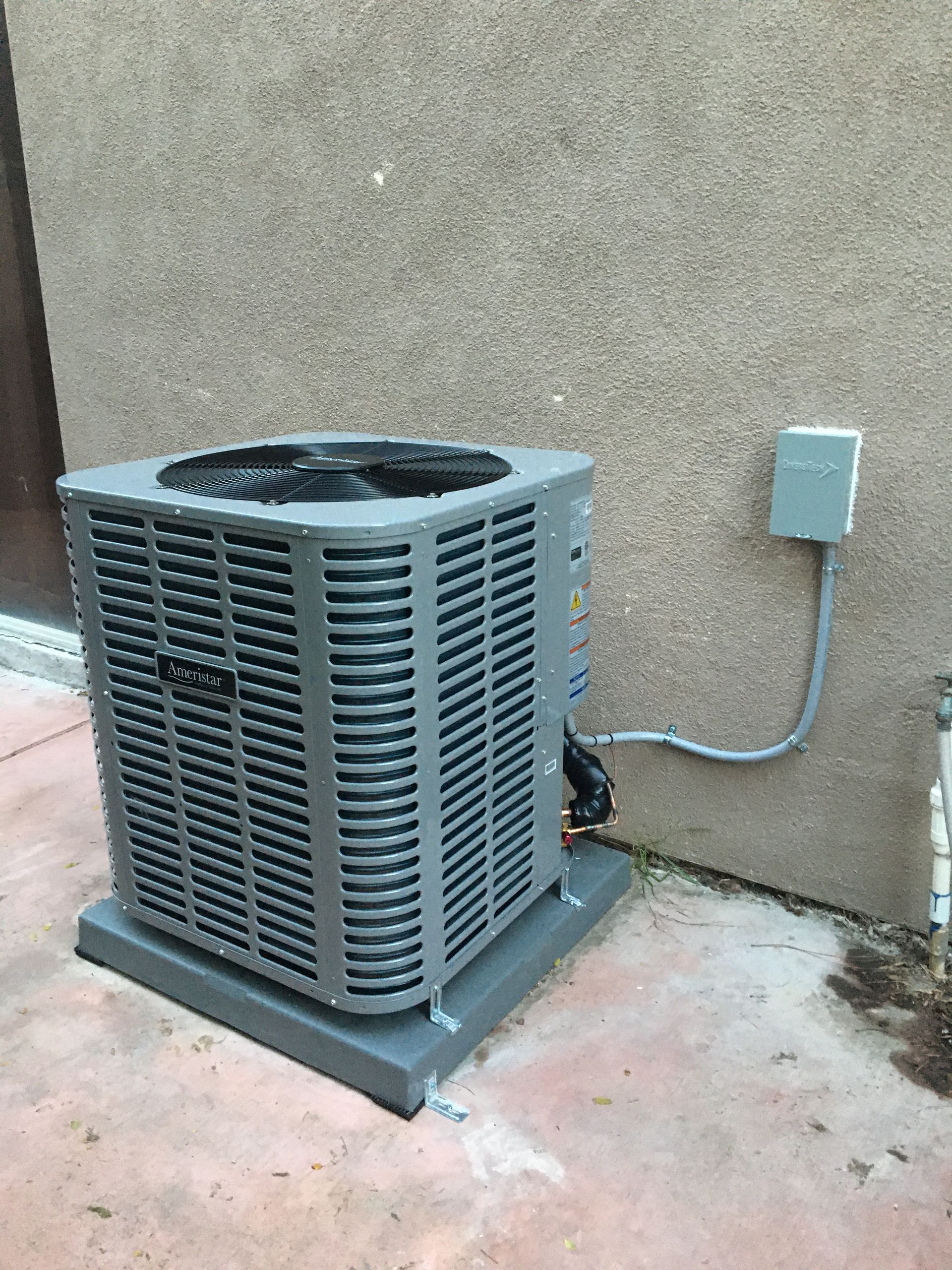 Outdoor air conditioner unit beside a beige wall with a conduit box and concrete pad.