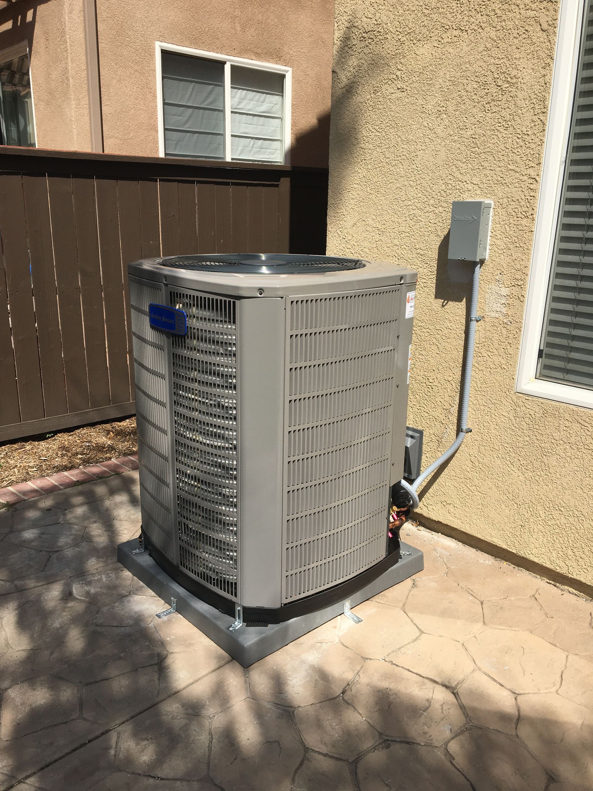 Outdoor air conditioner unit beside a stucco house wall and fence in a sunny backyard