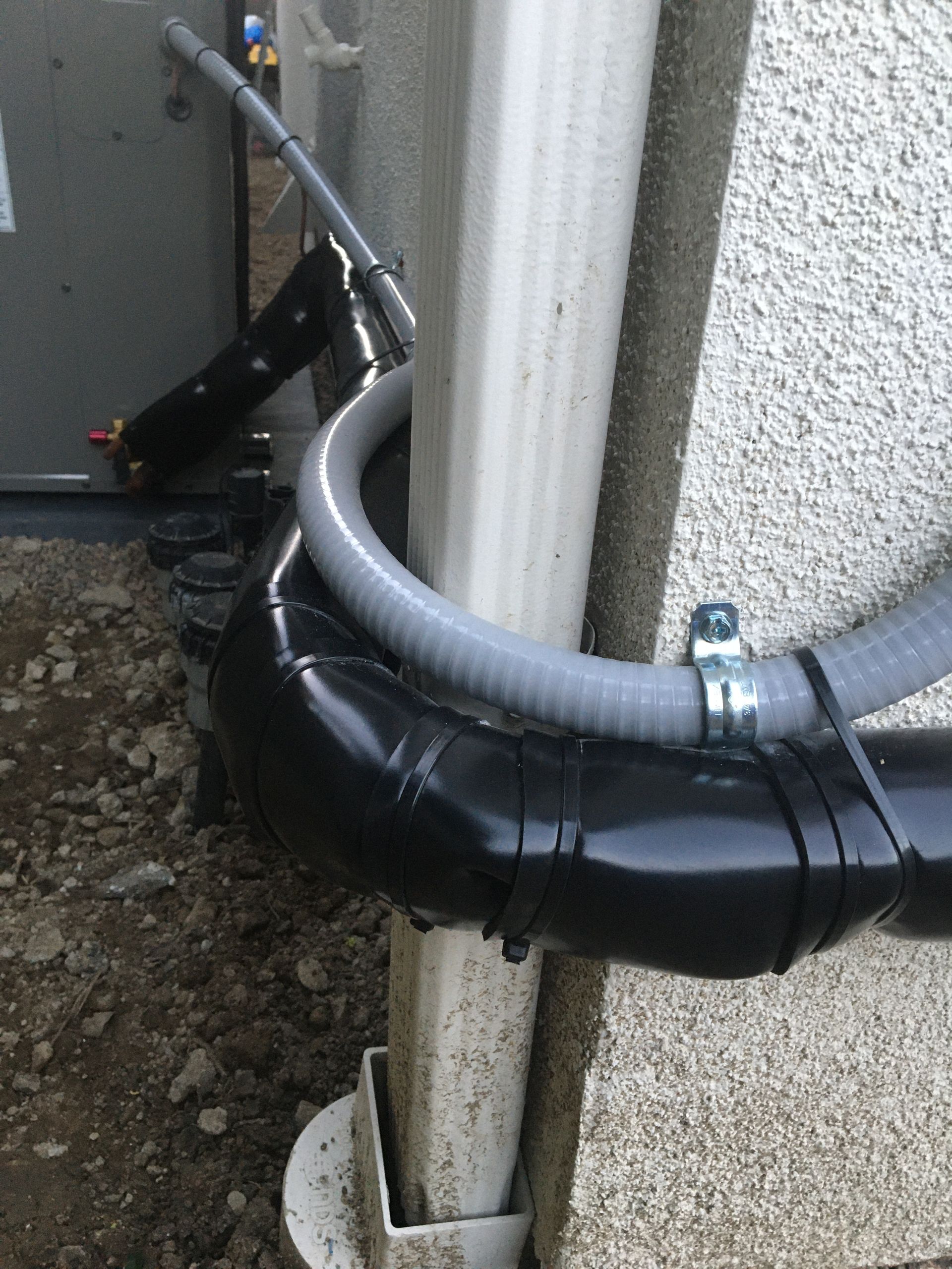 Black insulated pipe wrapped around a white vertical pipe beside a wall outdoors.