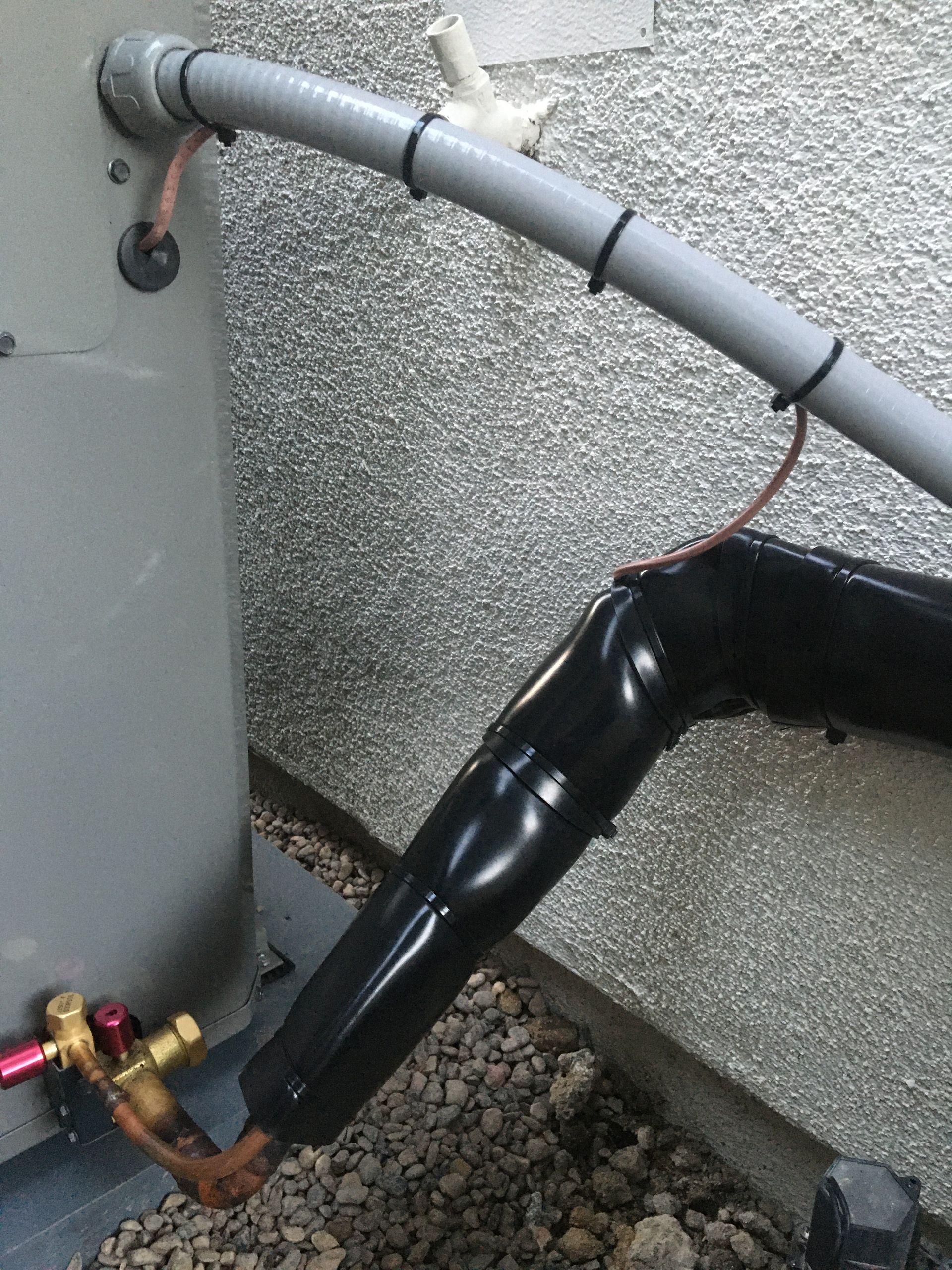 Black pipe wrapped in plastic connects to a wall-mounted gray pipe and brass fitting outdoors.