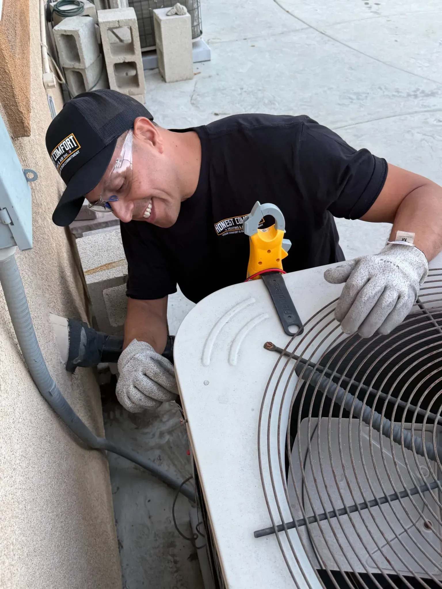 Technician in black cap and gloves works on an outdoor air conditioner unit with a yellow tool.