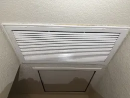 Ceiling vent with white grille above a rectangular attic access panel on a beige ceiling