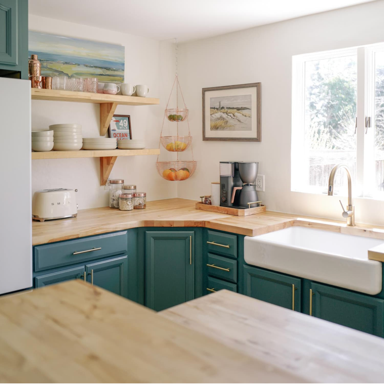 Bright kitchen with teal cabinets, wooden counters, open shelves, and a white farmhouse sink by a sunny window