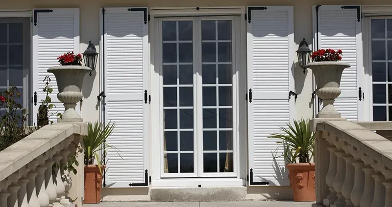 Portes-fenêtres blanches à volets, encadrées de plantes en pot sur un balcon ensoleillé