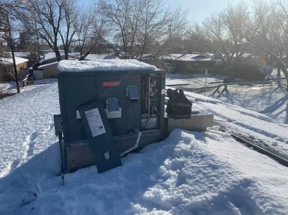 A grey HVAC rooftop unit with an open service panel sits on a snowy roof with a residential neighborhood in the background.