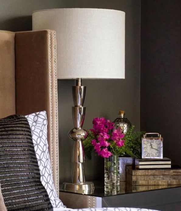A lamp sits on a nightstand next to a vase of pink flowers and a clock
