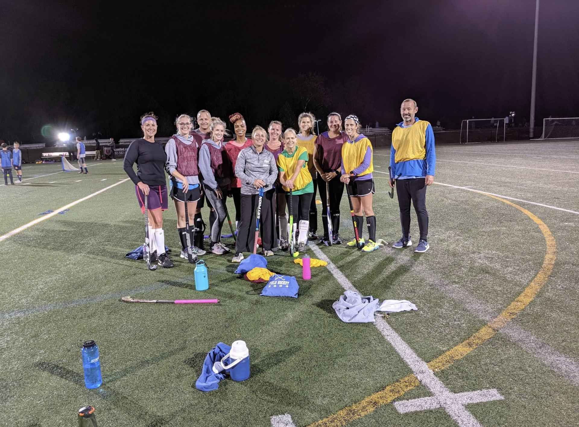 Group of people posing on a lit soccer field at night with sports gear laid out in front of them