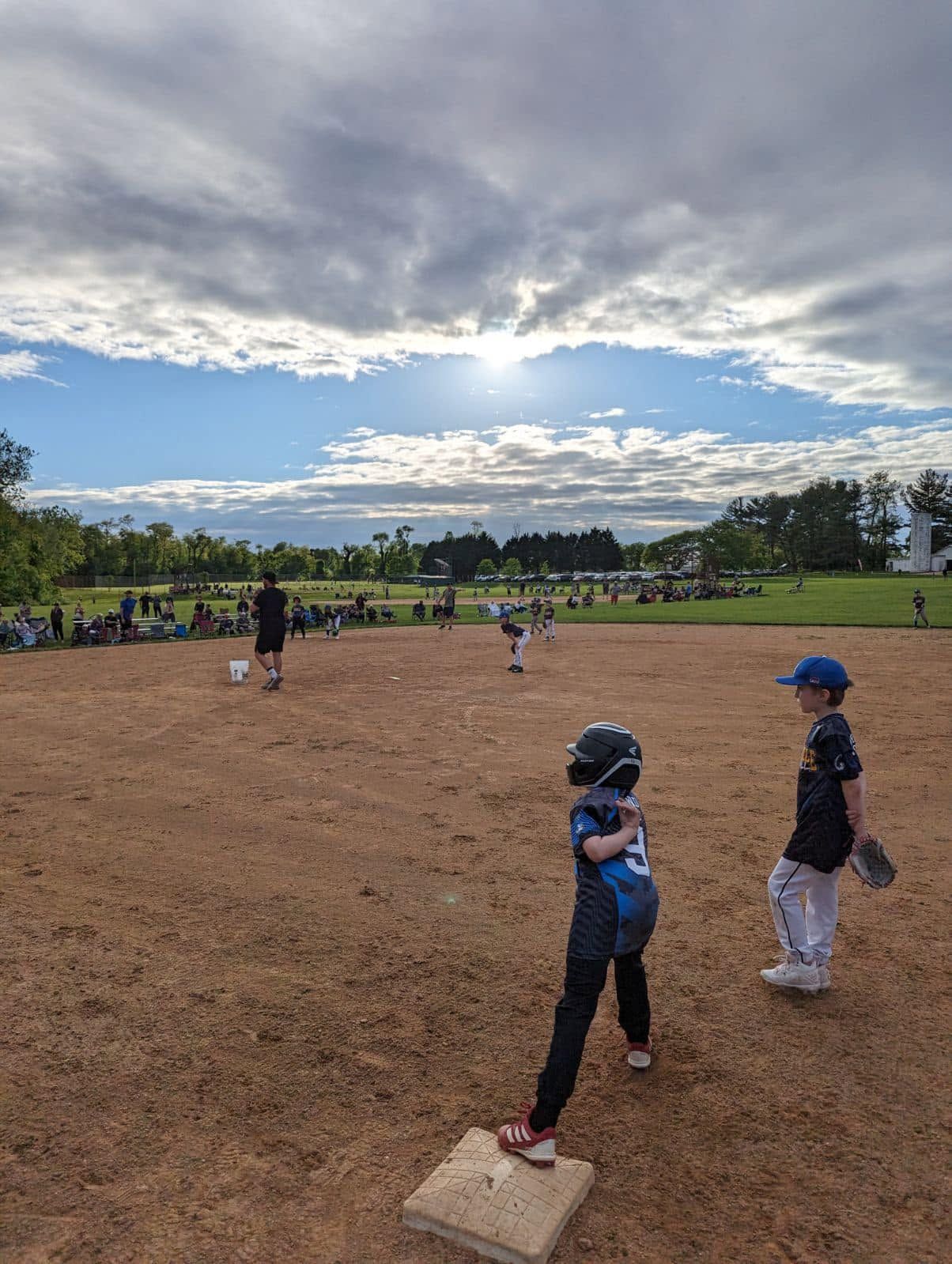 Children playing baseball on a dirt field under a cloudy sky, with one child in a blue cap in the foreground