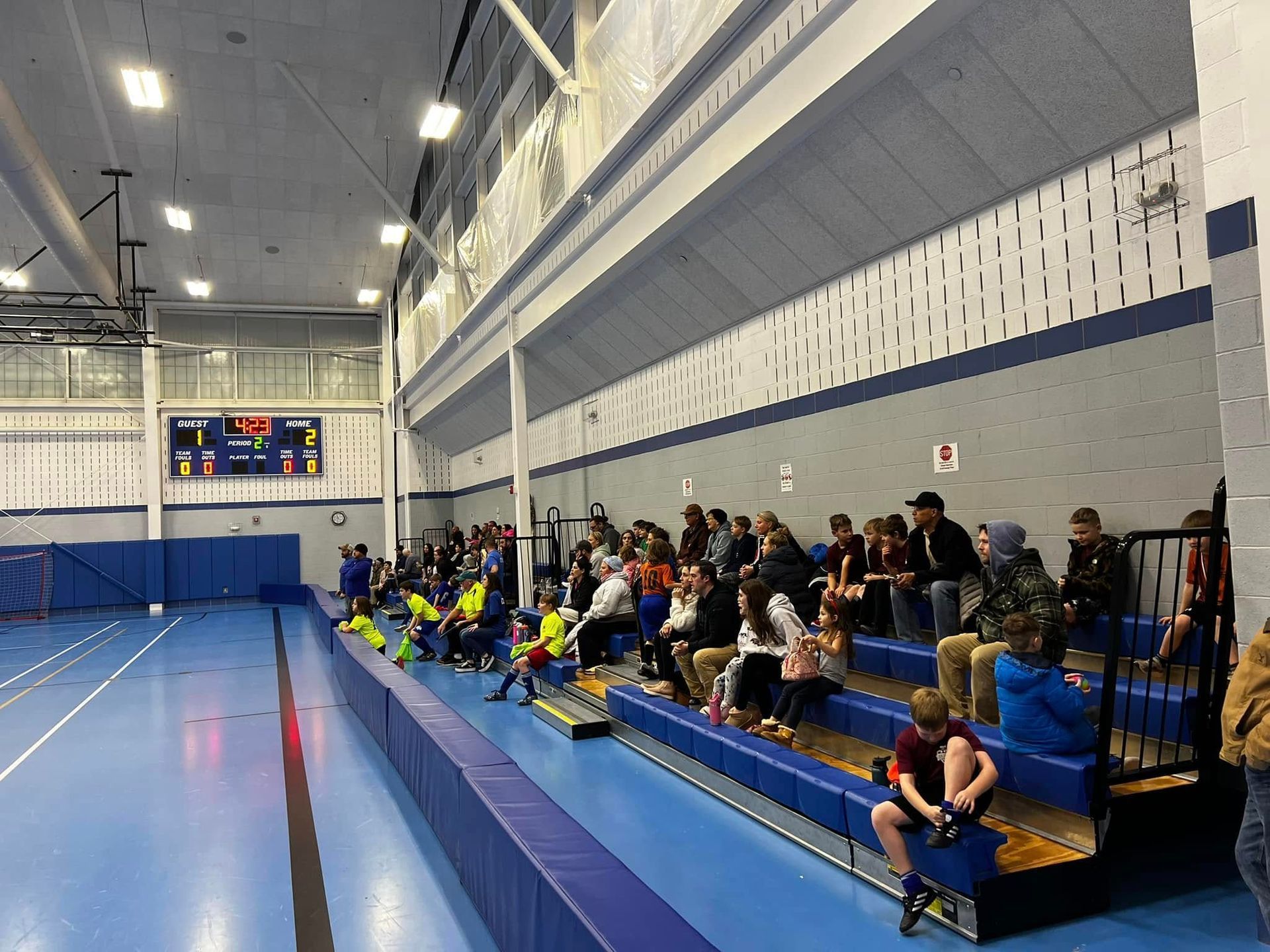 Spectators seated along the bleachers in an indoor gymnasium beside a blue sports floor