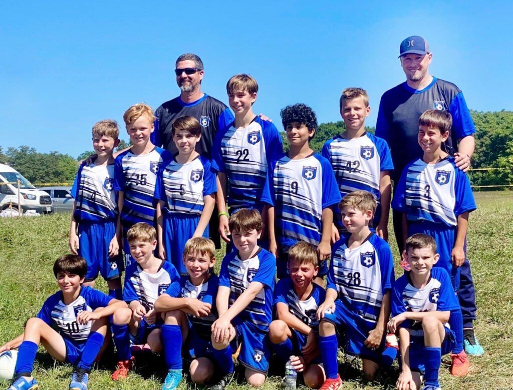 Youth soccer team in blue jerseys posing with coaches outdoors on a sunny field.