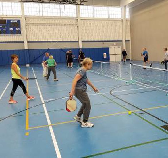 Indoor gym with several people playing pickleball on blue courts using paddles and a ball