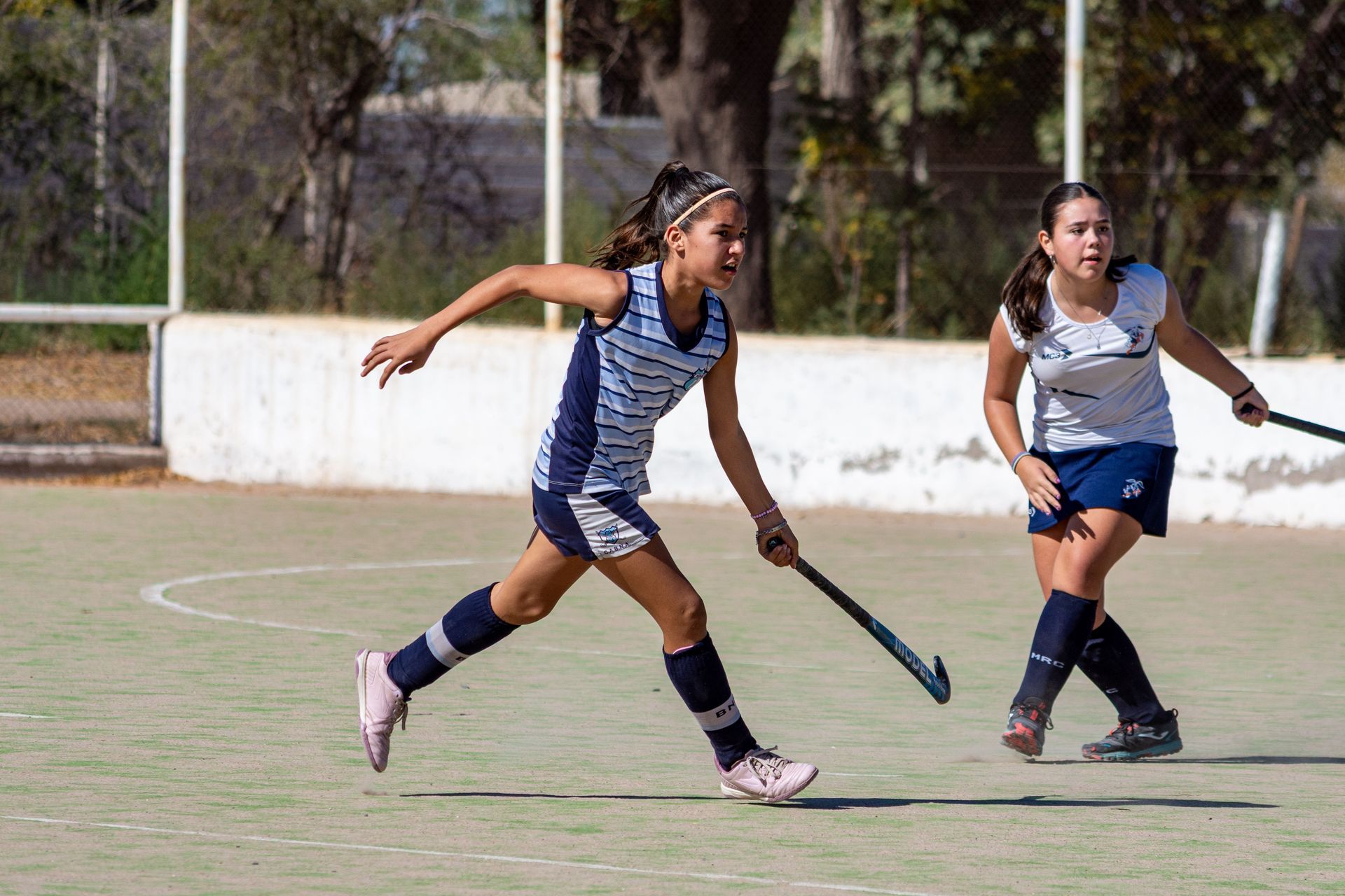 Two field hockey players in blue uniforms chasing the ball on a grassy field