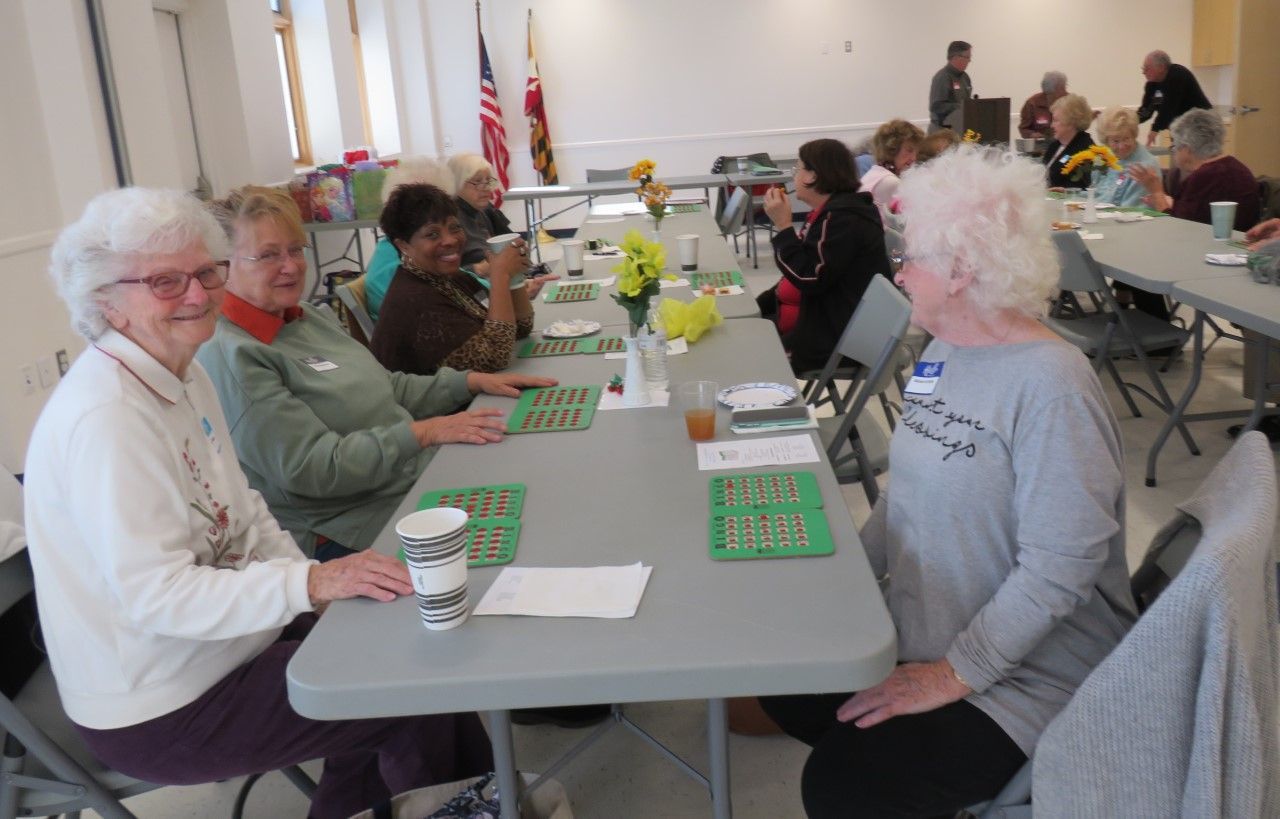 People seated at tables in a bright hall, playing a board game and chatting