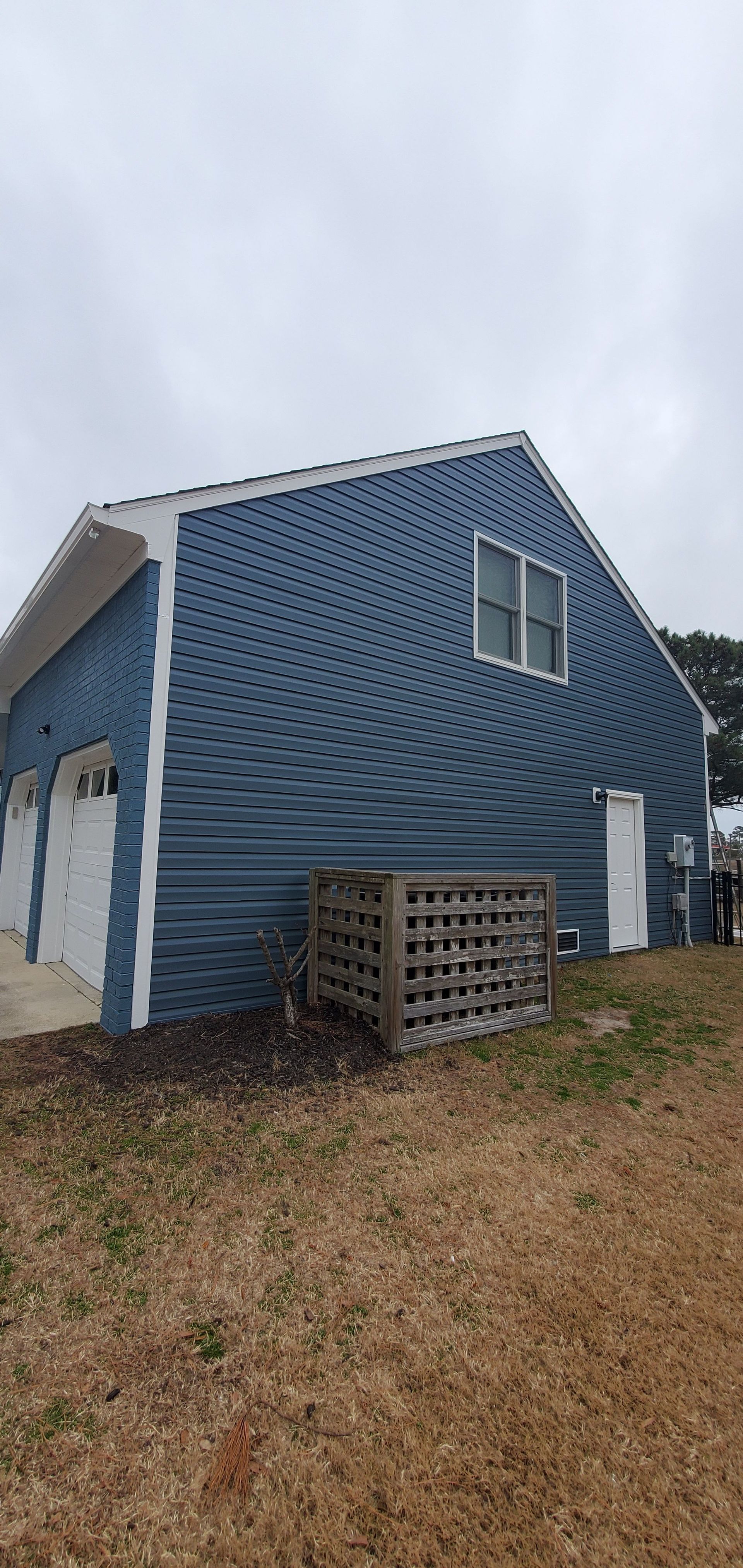 Blue vinyl-sided house with white trim and windows on a grassy slope under cloudy sky