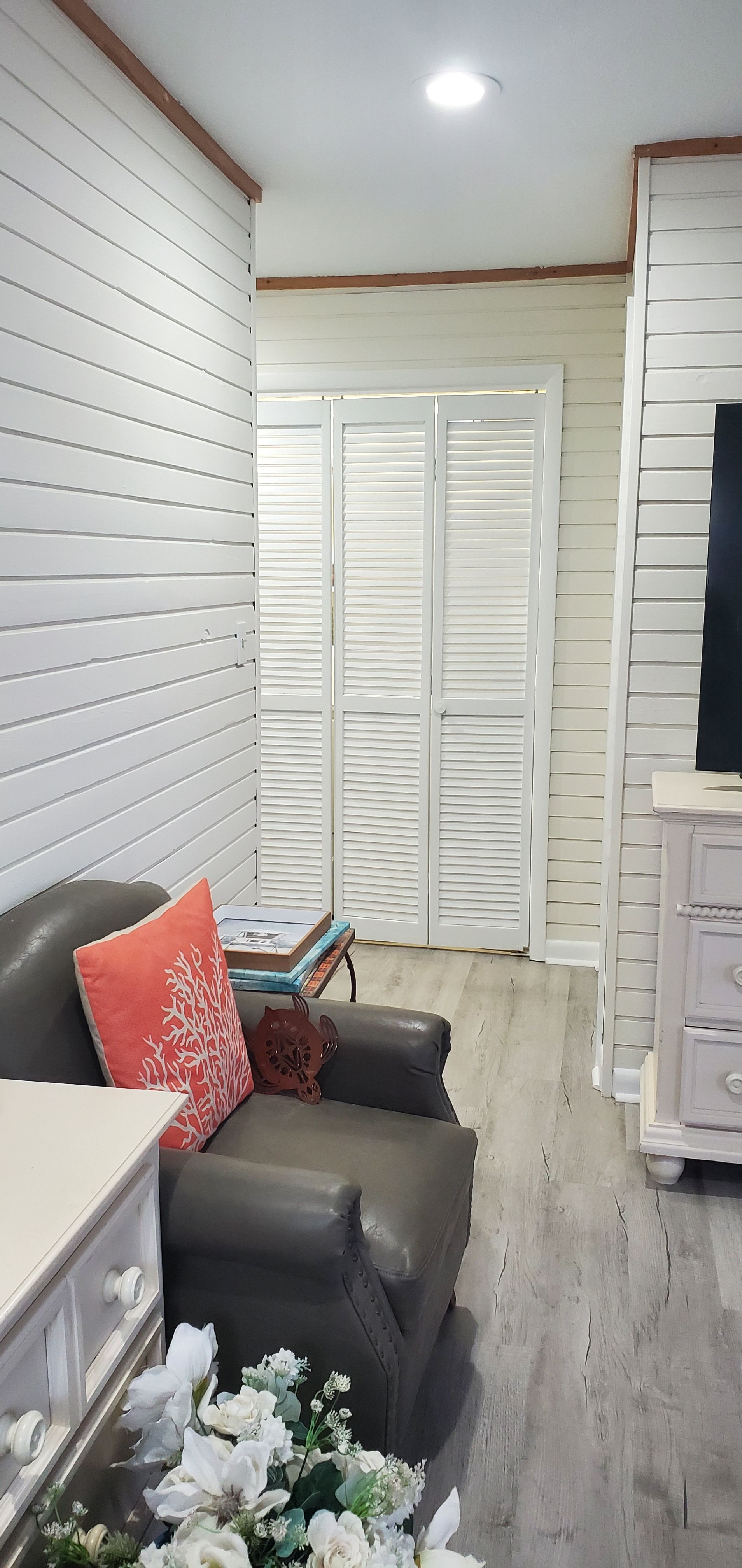 Bright narrow living room with white paneled walls, gray sofa, pink pillow, and louvered closet doors.