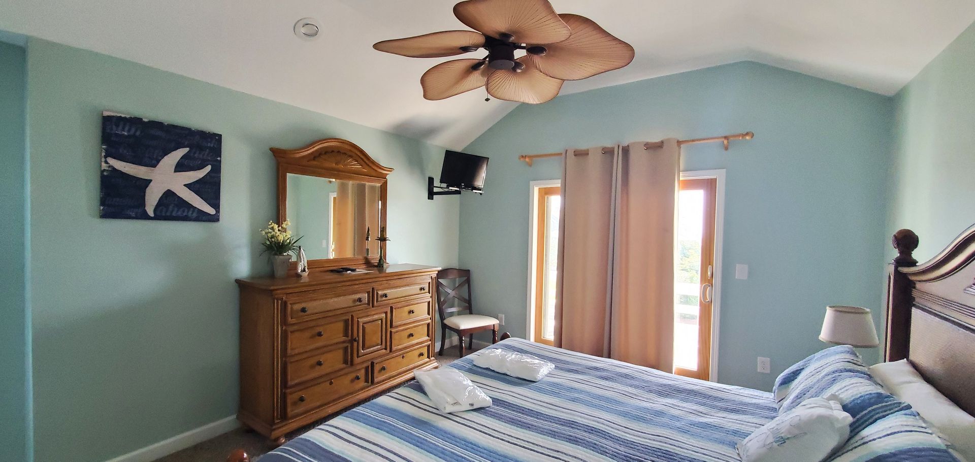 Bright bedroom with blue walls, a striped bed, wooden dresser, ceiling fan, and curtains over a window.
