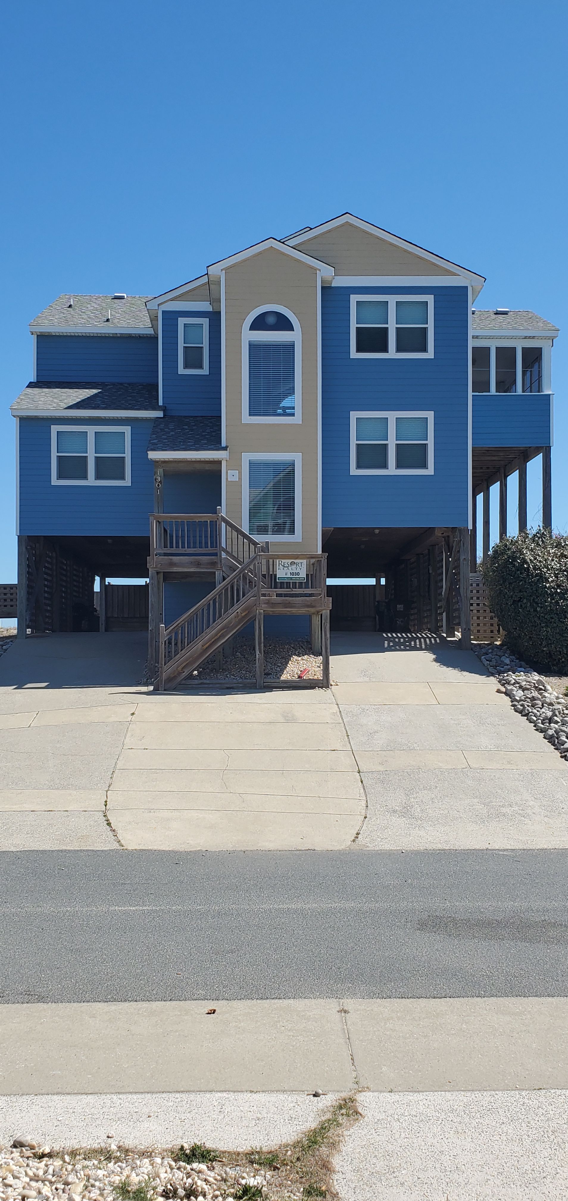 Blue two-story beach house with a front staircase, porch, and parking area under a clear sky