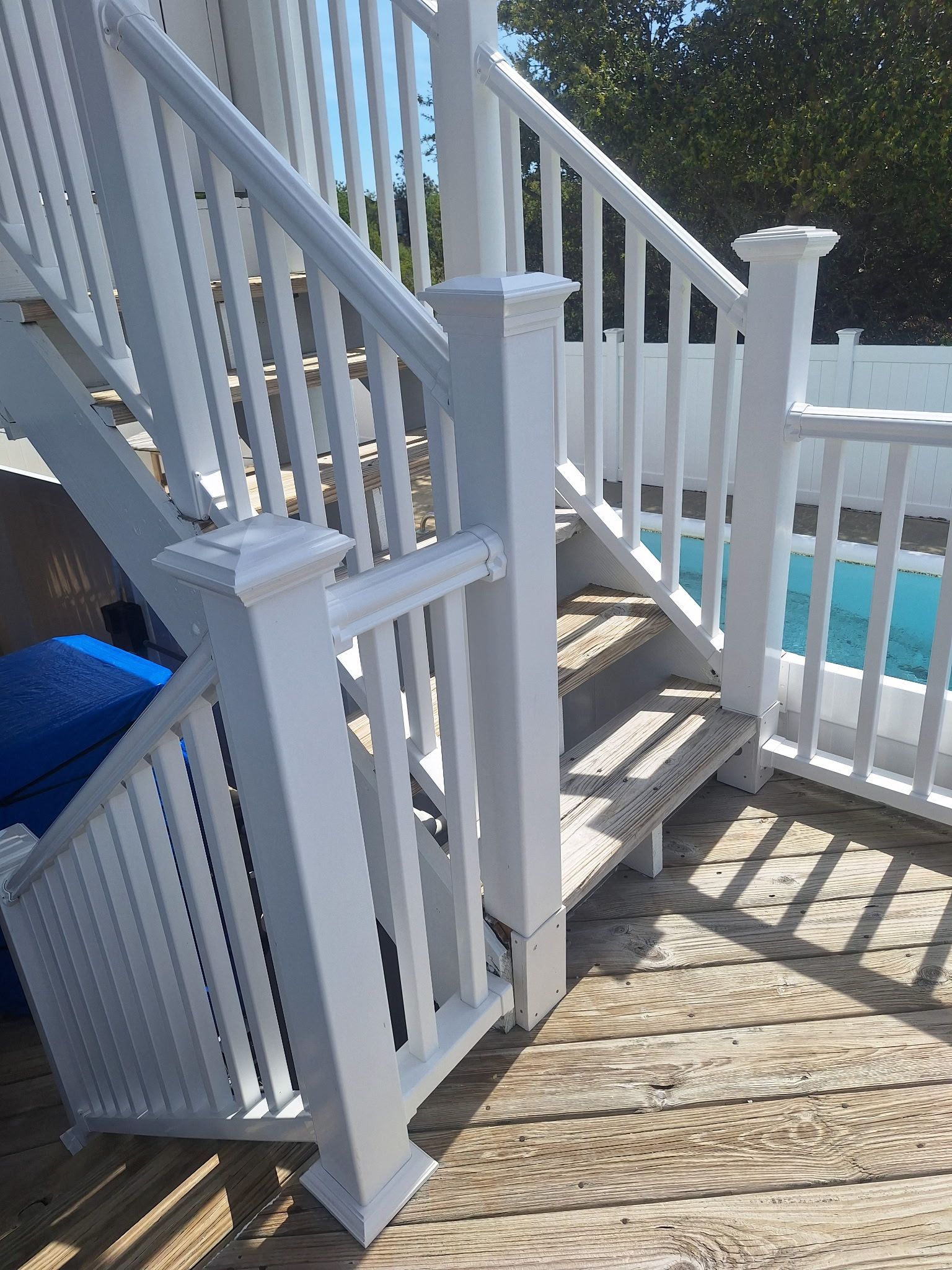 White deck stairs with railing beside a blue pool and gate, viewed from a wooden porch