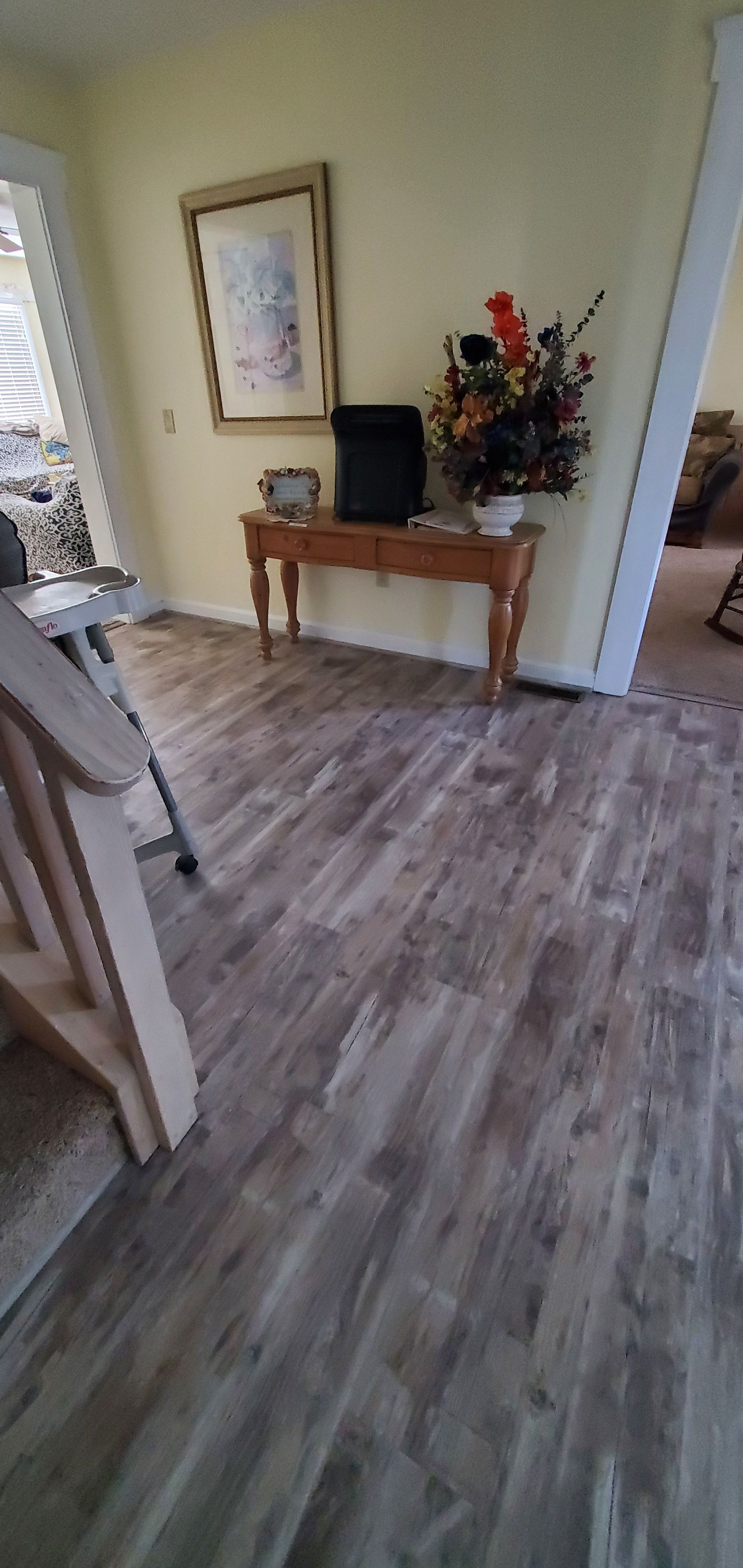 Hallway with gray wood floors, small wooden table, framed art, and flowers near an open doorway