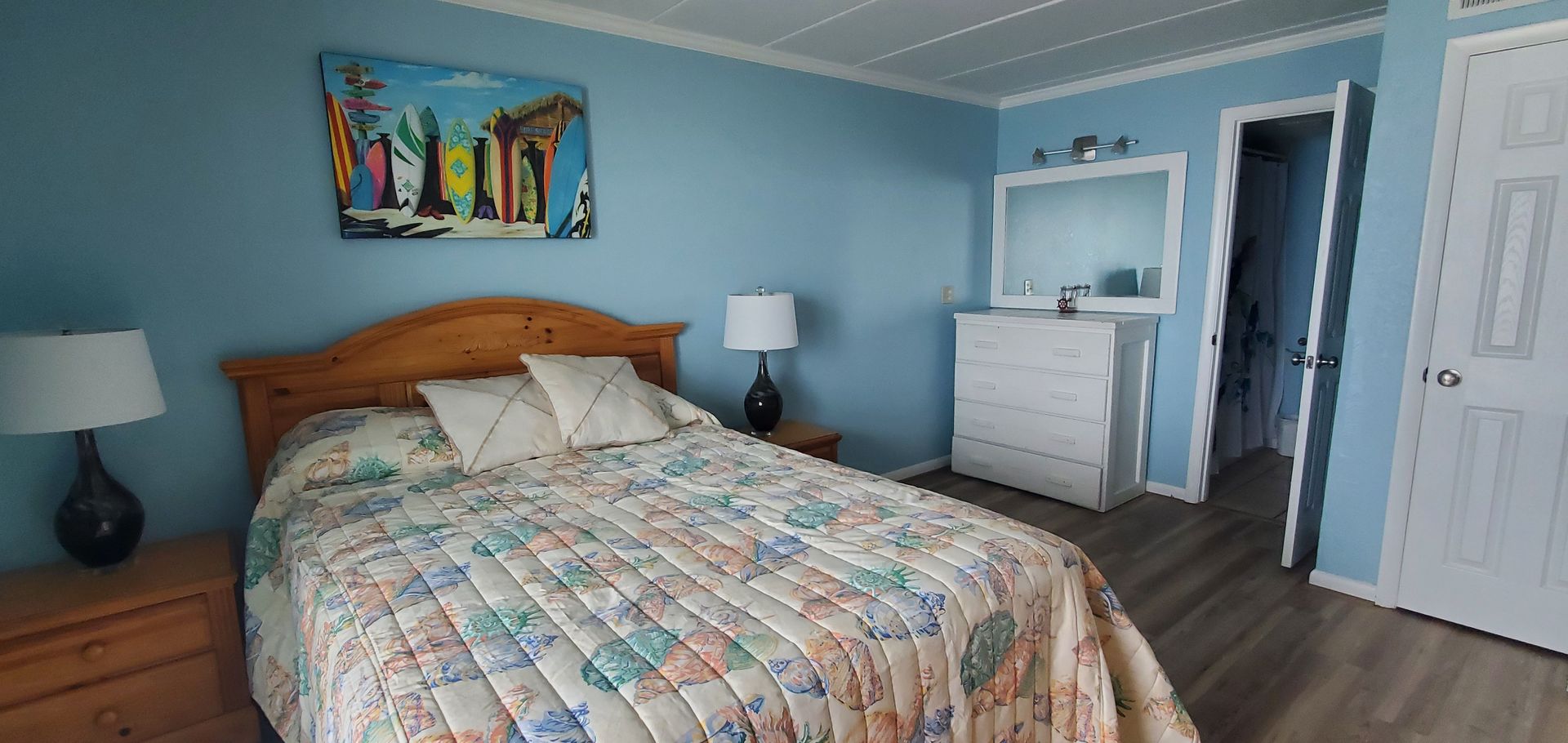 Blue bedroom with a bed, dresser, two lamps, and an open doorway to a bathroom.