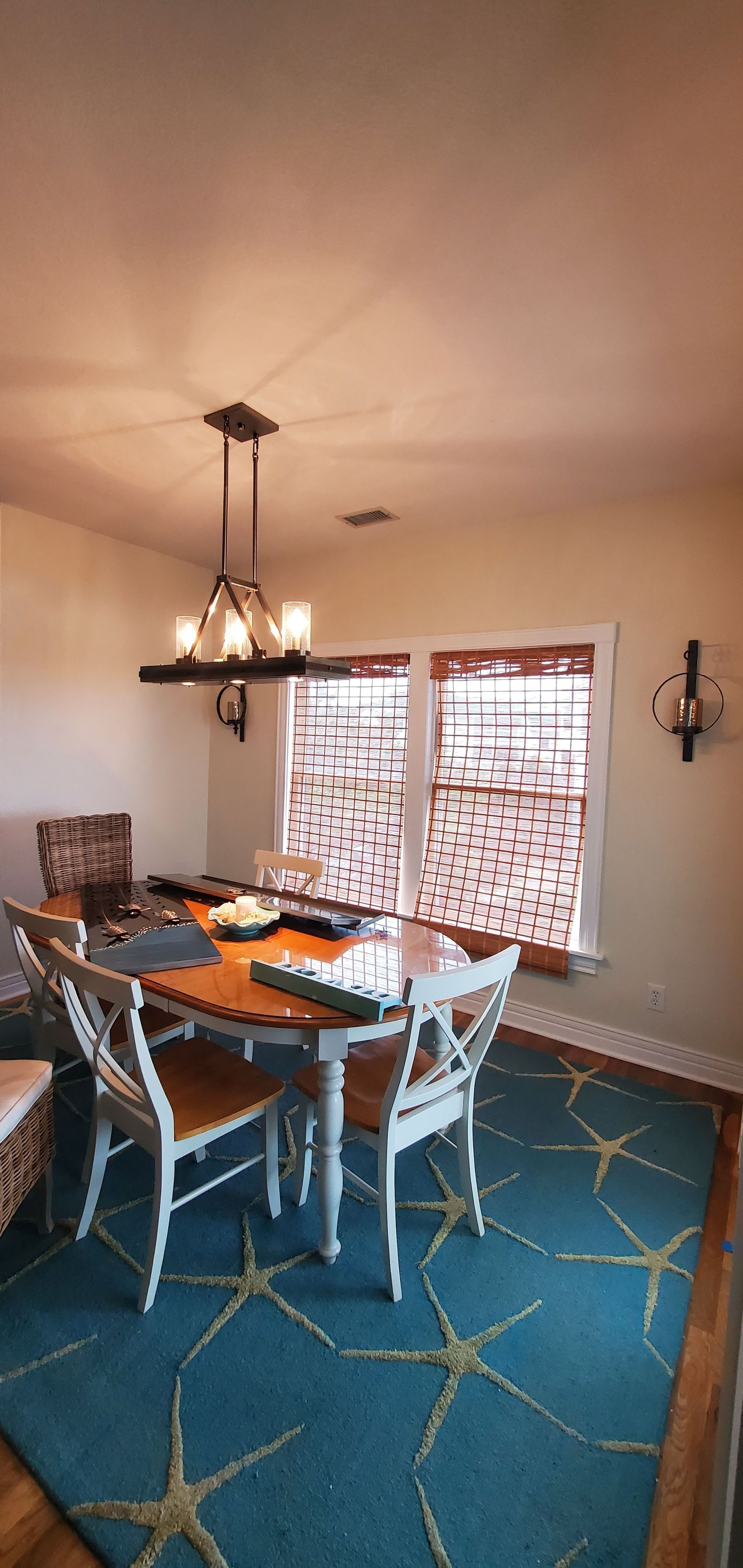 Dining room with blue rug, glass table, white chairs, and chandelier near a large window with wood blinds