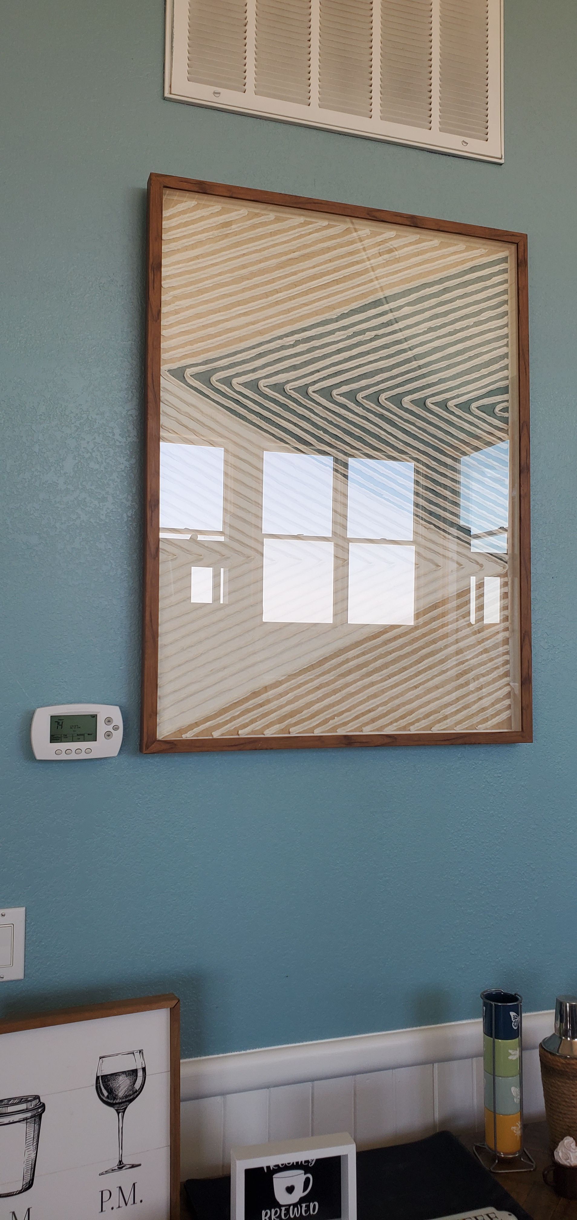 Mirror on a teal wall above a small shelf with glassware and bottles, reflecting window light and a ceiling grate