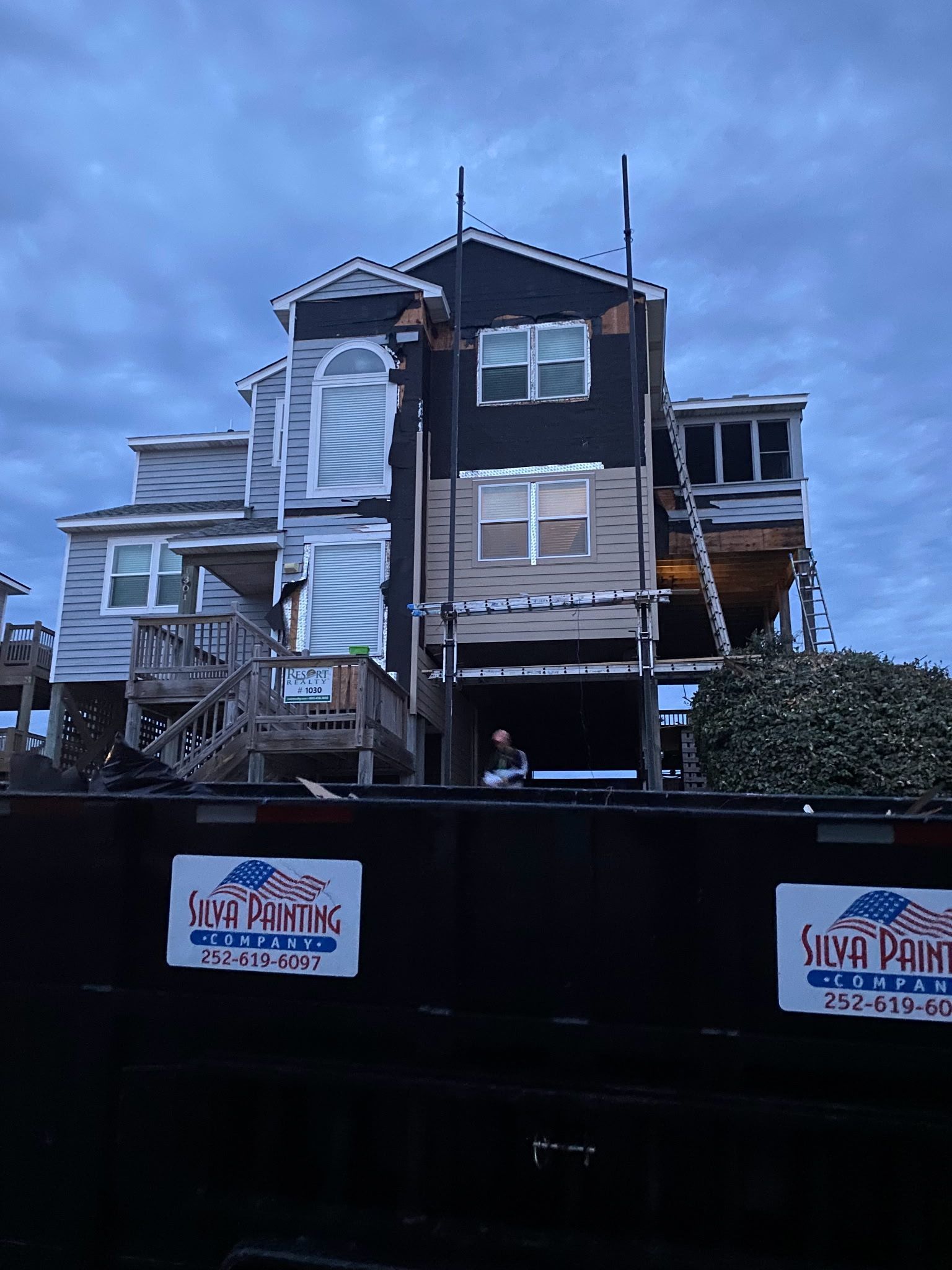 Damaged multi-story house behind a black fence, with a blue sky and a construction sign in front.