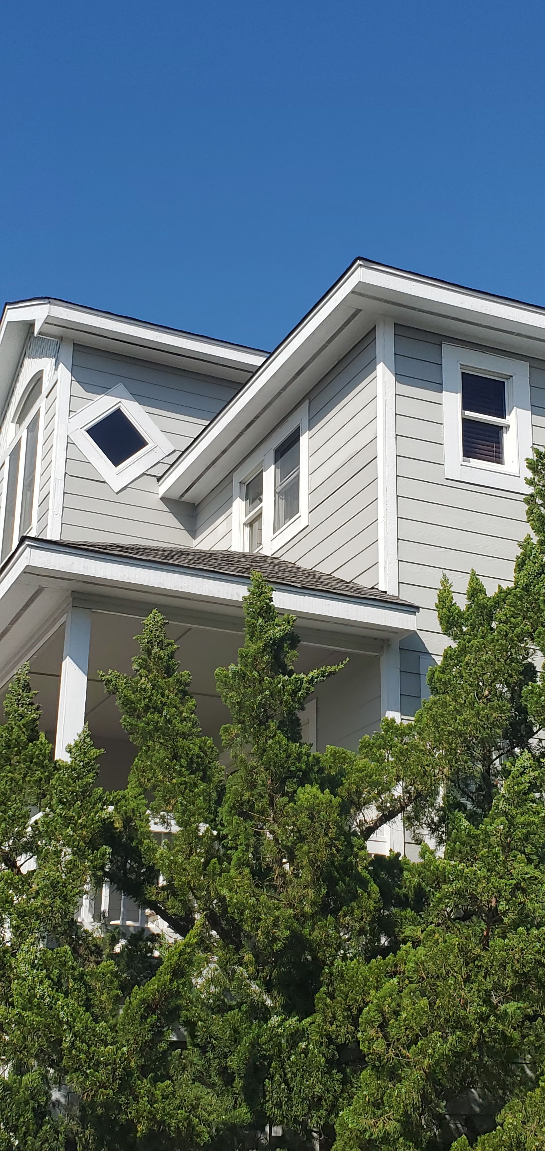 White house with gray roof and dormer windows, partly hidden by green trees under a bright sky