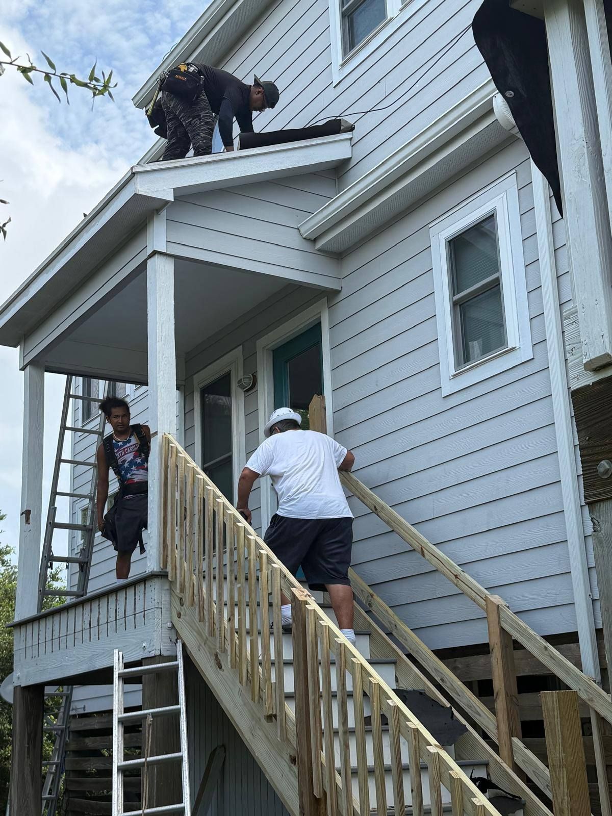 Workers on a house roof and porch, repairing gray siding and a wooden staircase.