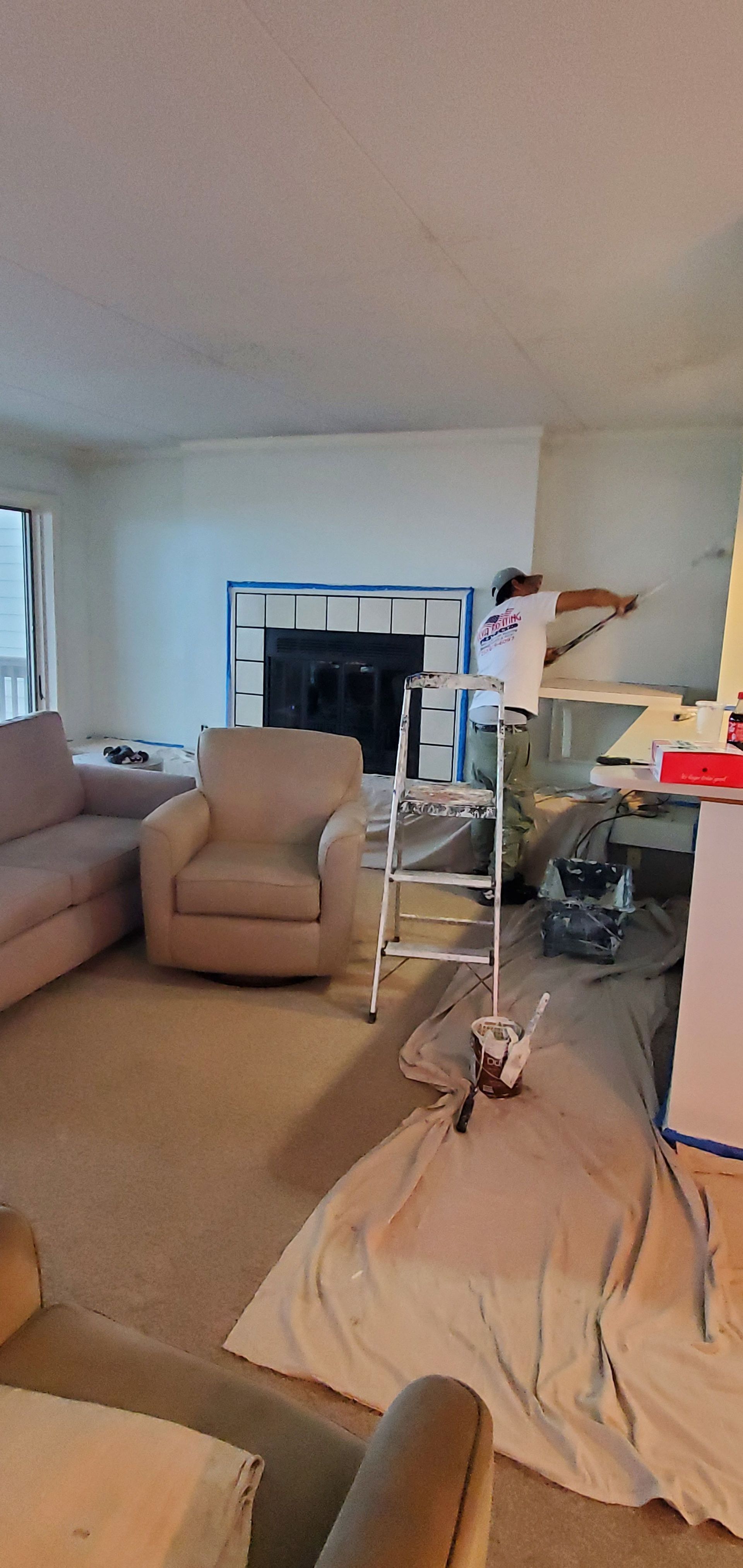 Cluttered living room with a beige recliner, white tarp on the floor, and tools near a fireplace and window.