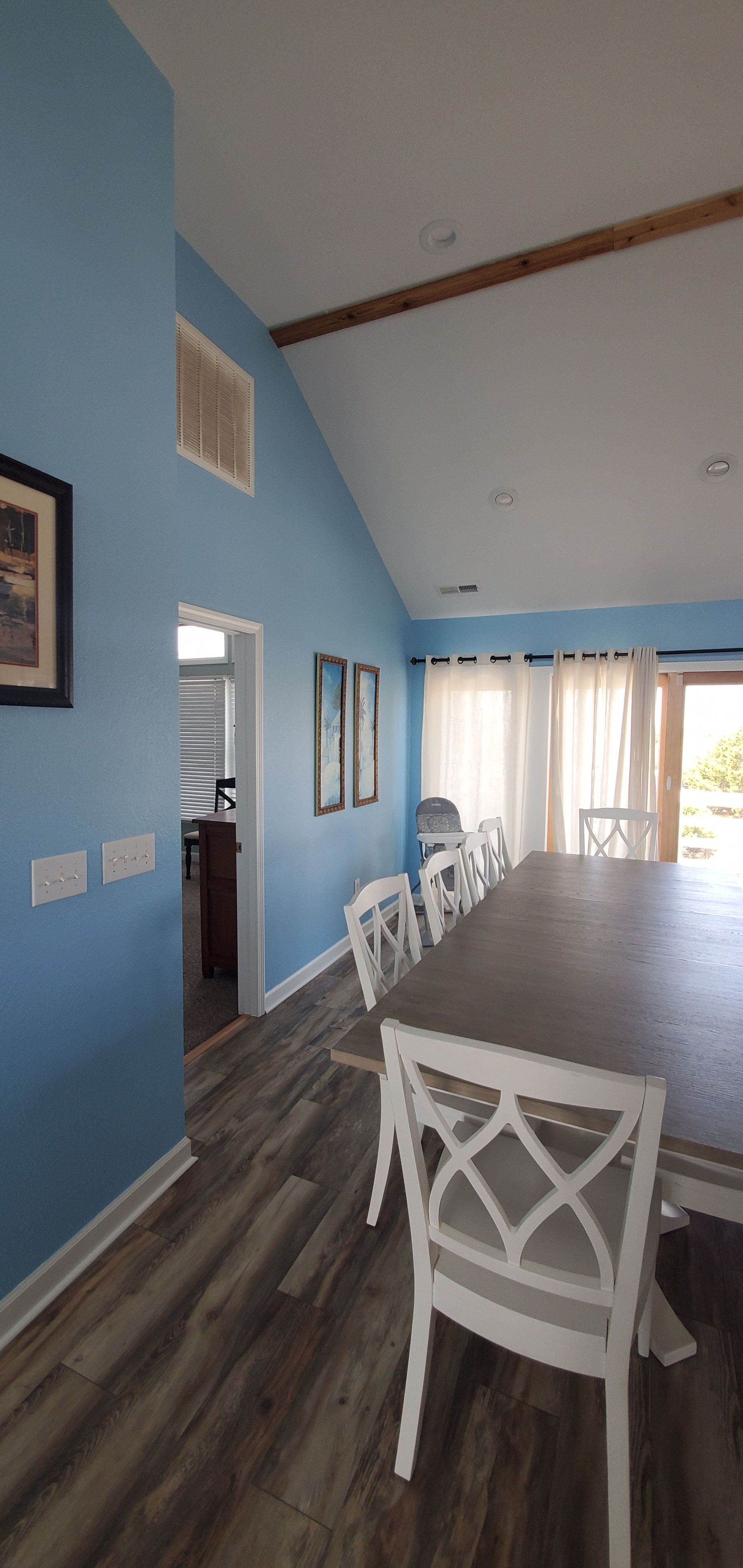 Blue dining room with white chairs and a long wooden table beside bright windows and a sliding glass door