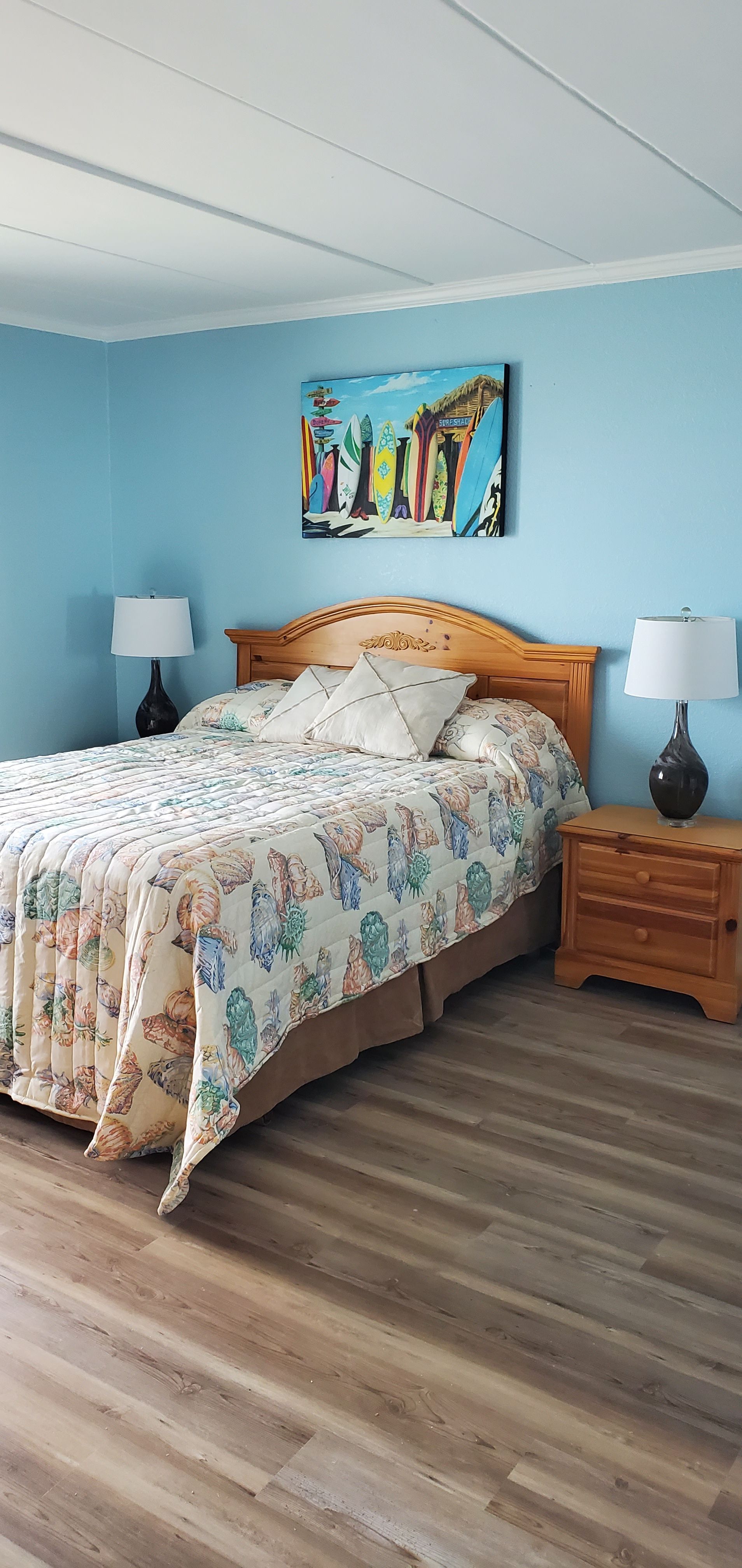 Light blue bedroom with a wooden bed, floral bedding, twin lamps, and colorful wall art