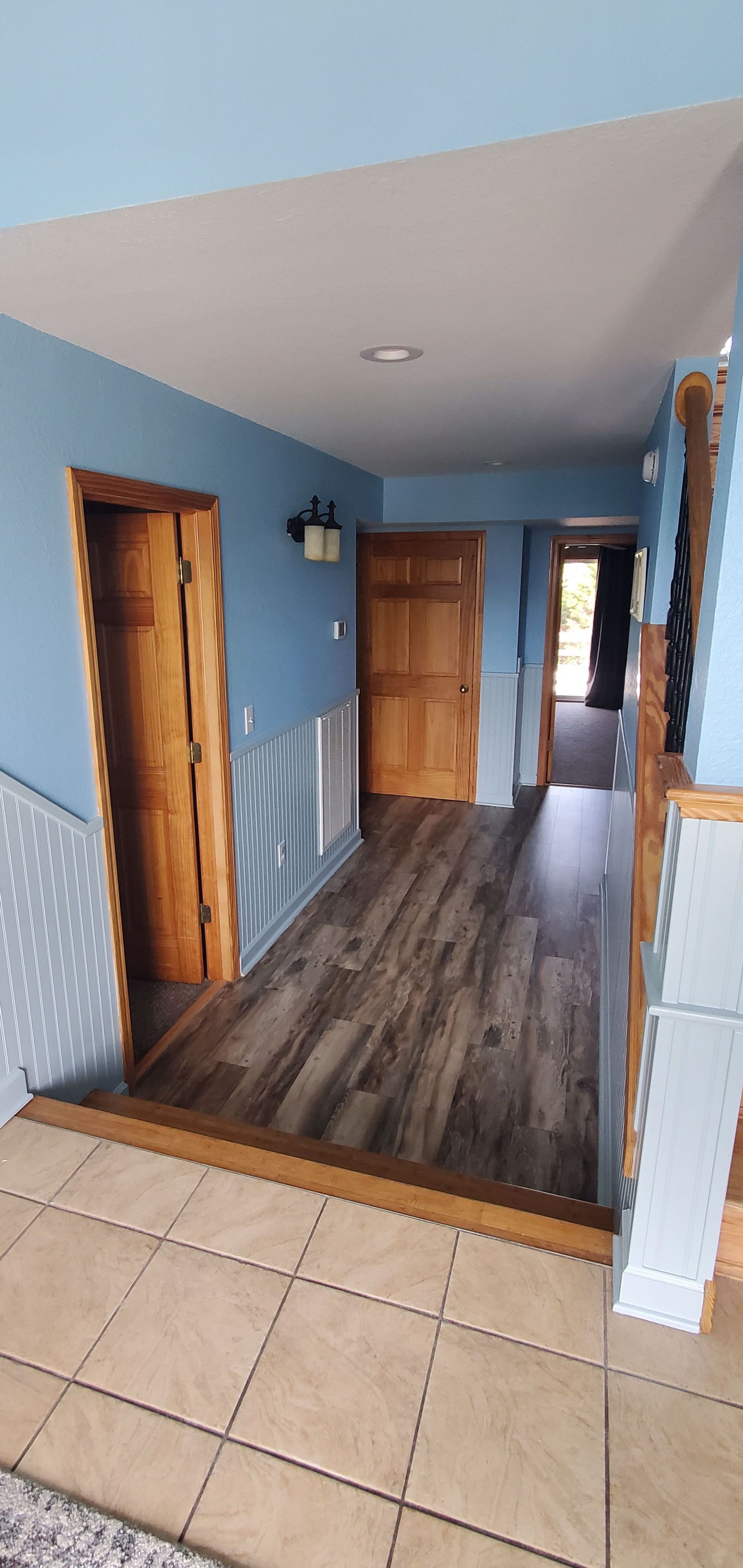 Narrow hallway with blue walls, wood doors, and tan tile leading to a dark wood floor.
