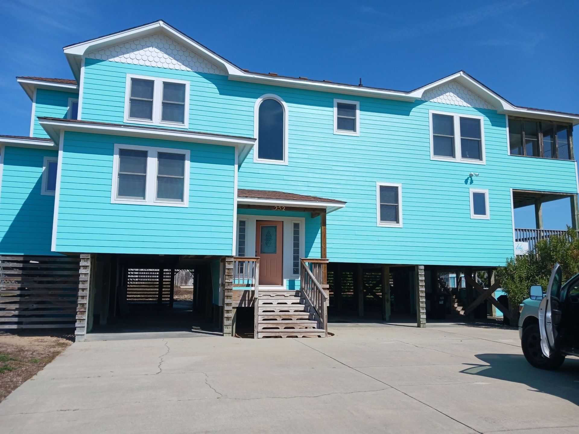 Elevated turquoise house on stilts with stairs and a driveway under a clear blue sky