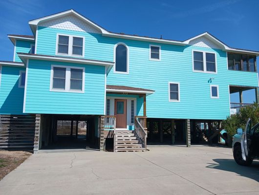 Bright turquoise beach house on stilts with front stairs and a driveway under a blue sky