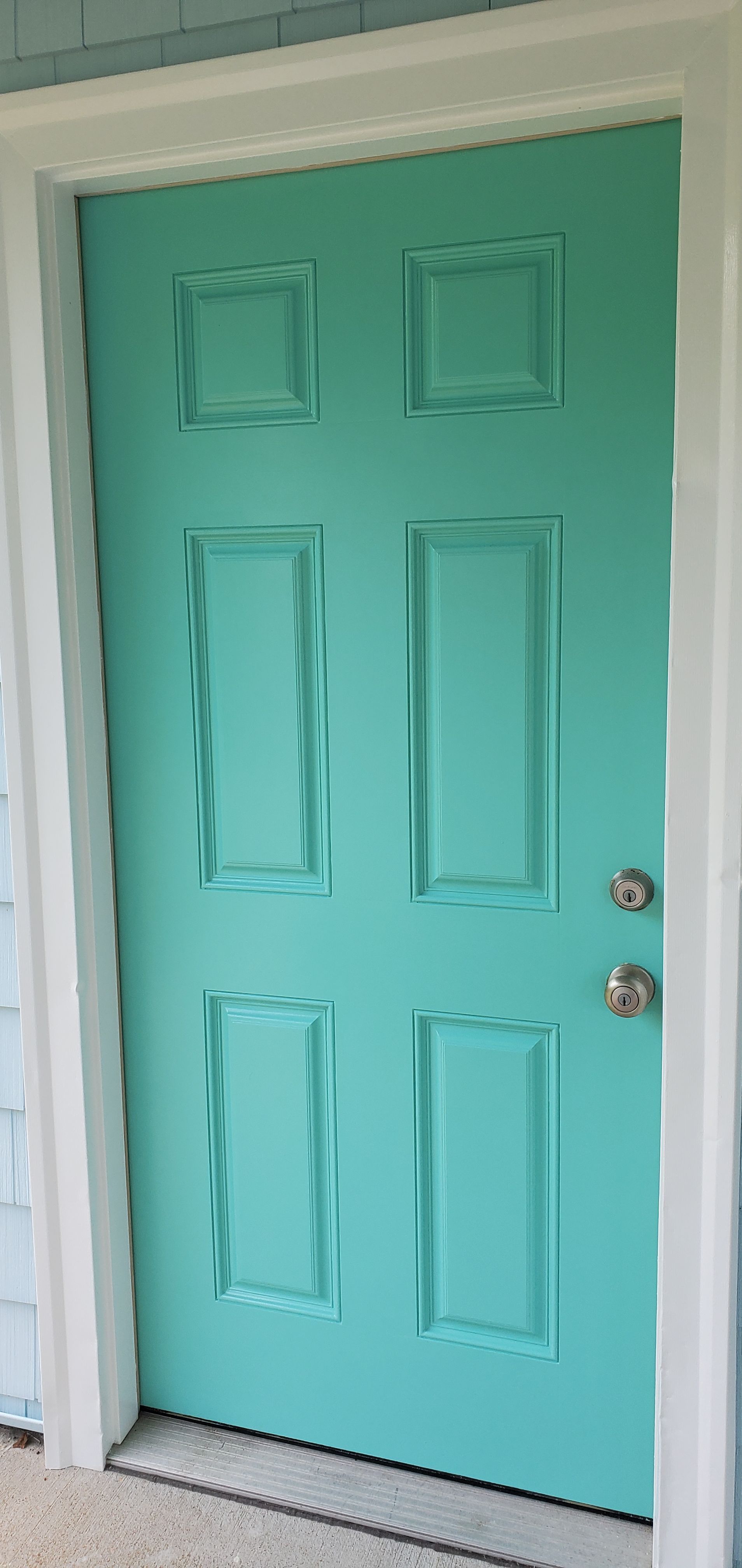 Turquoise front door with six raised panels and silver knob, framed by white trim and porch siding