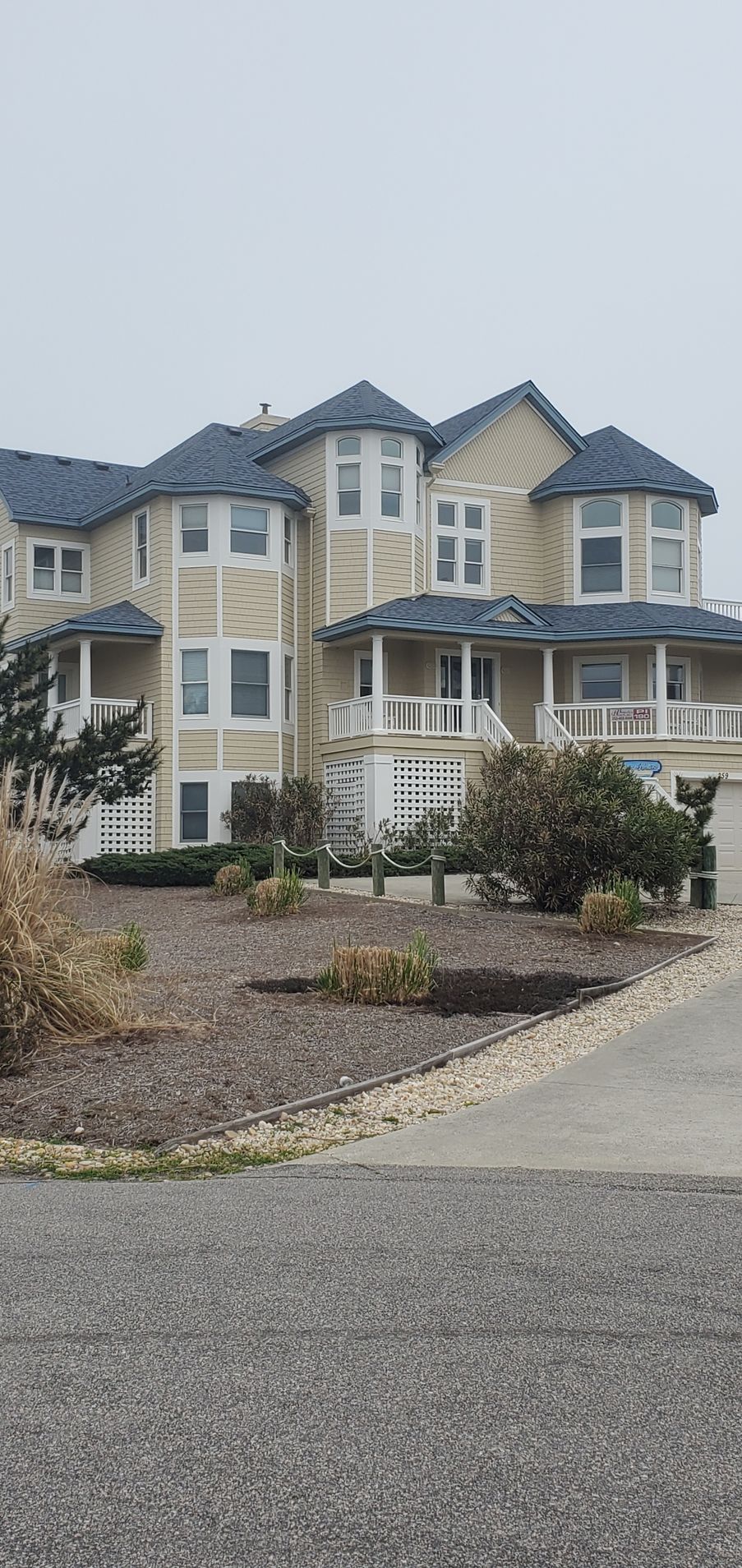 Large beige two-story house with blue-gray roof, porch, and landscaped front yard behind a gravel drive