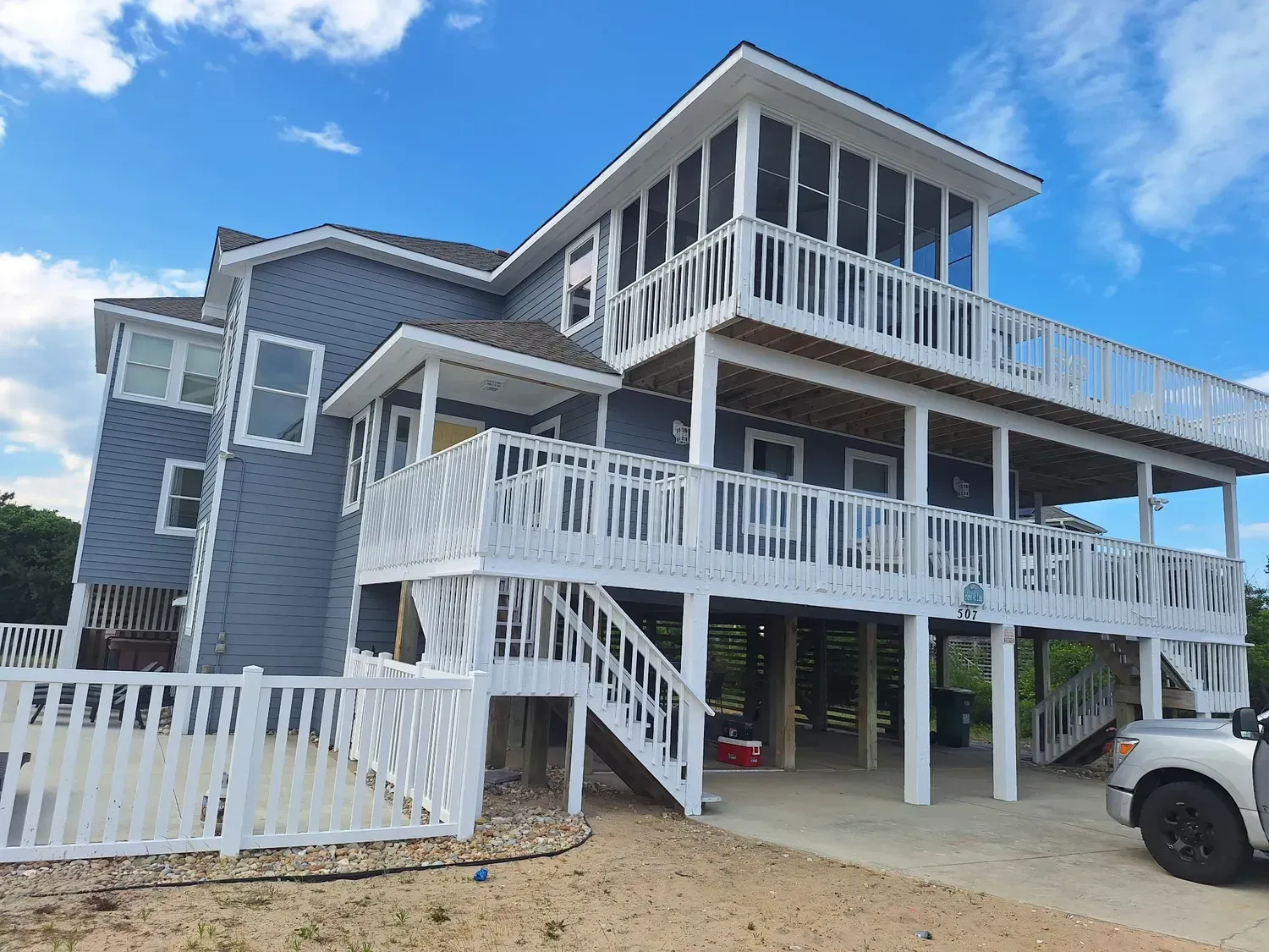 Gray beach house with white wraparound decks and stairs, under a blue sky with a parked white truck.