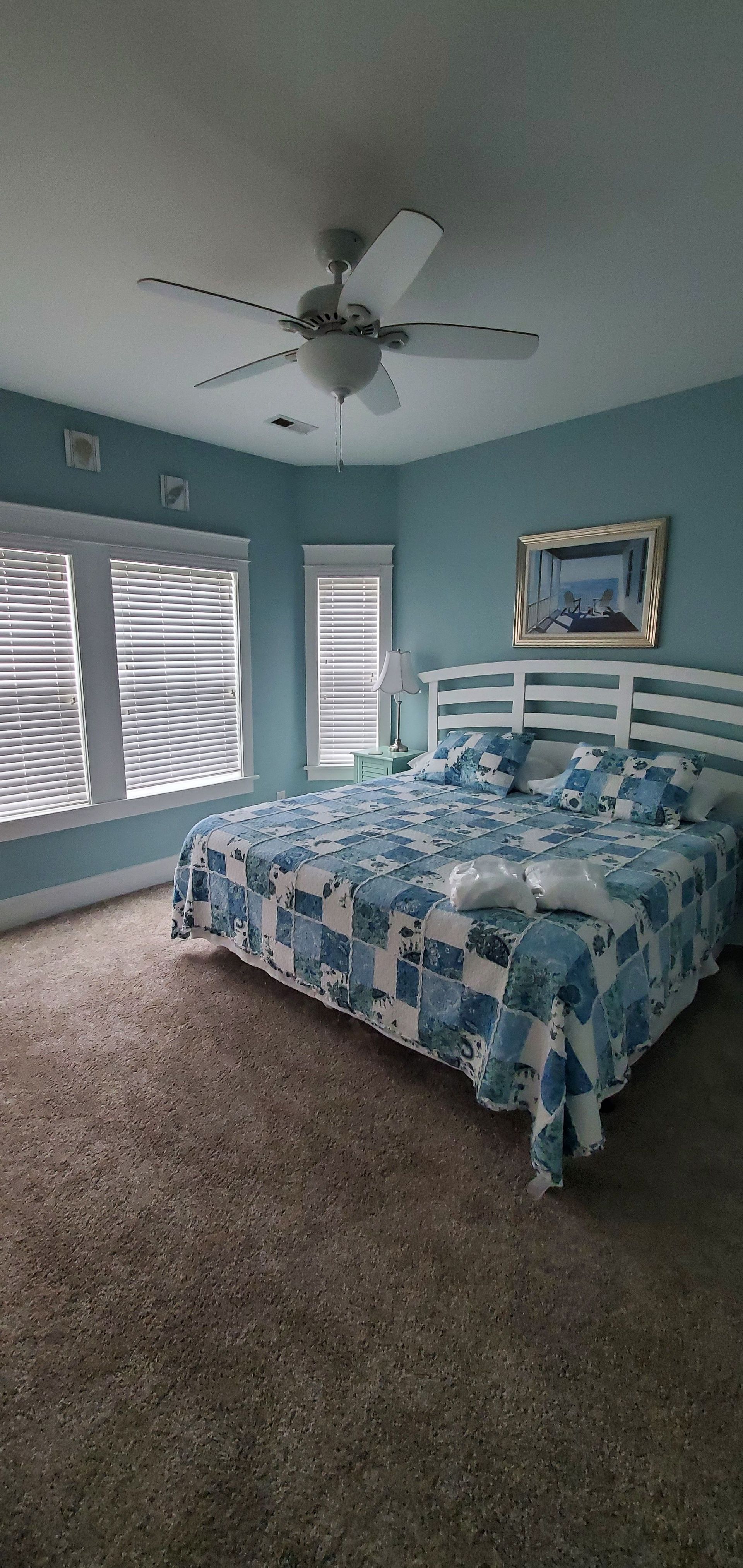 Bright bedroom with blue walls, patterned bedspread, ceiling fan, and ocean-view window curtains