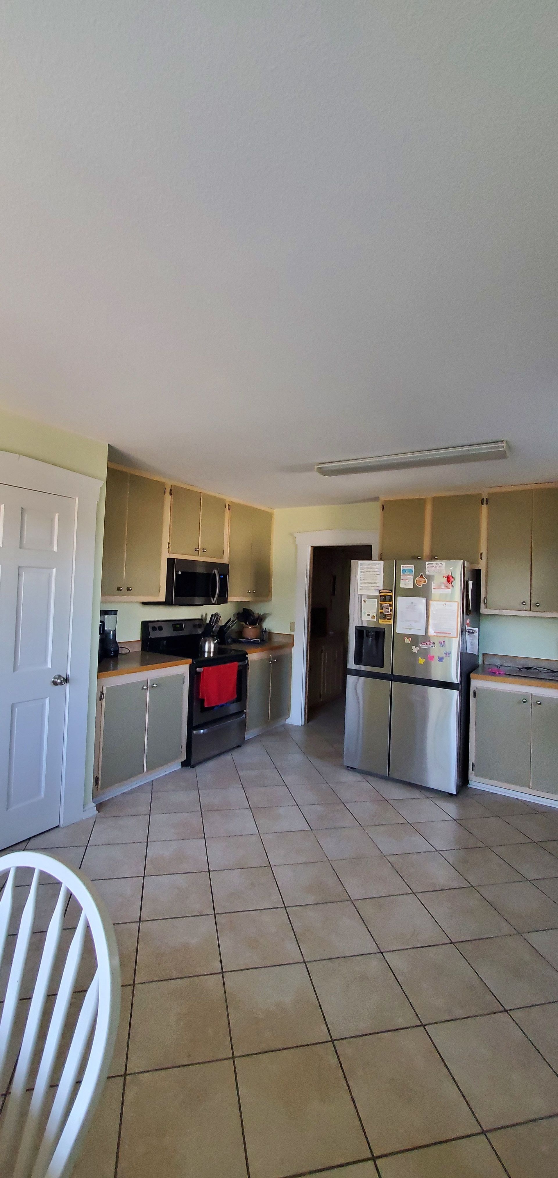 Bright kitchen with beige tile floor, white cabinets, stainless fridge, and an open doorway to another room.