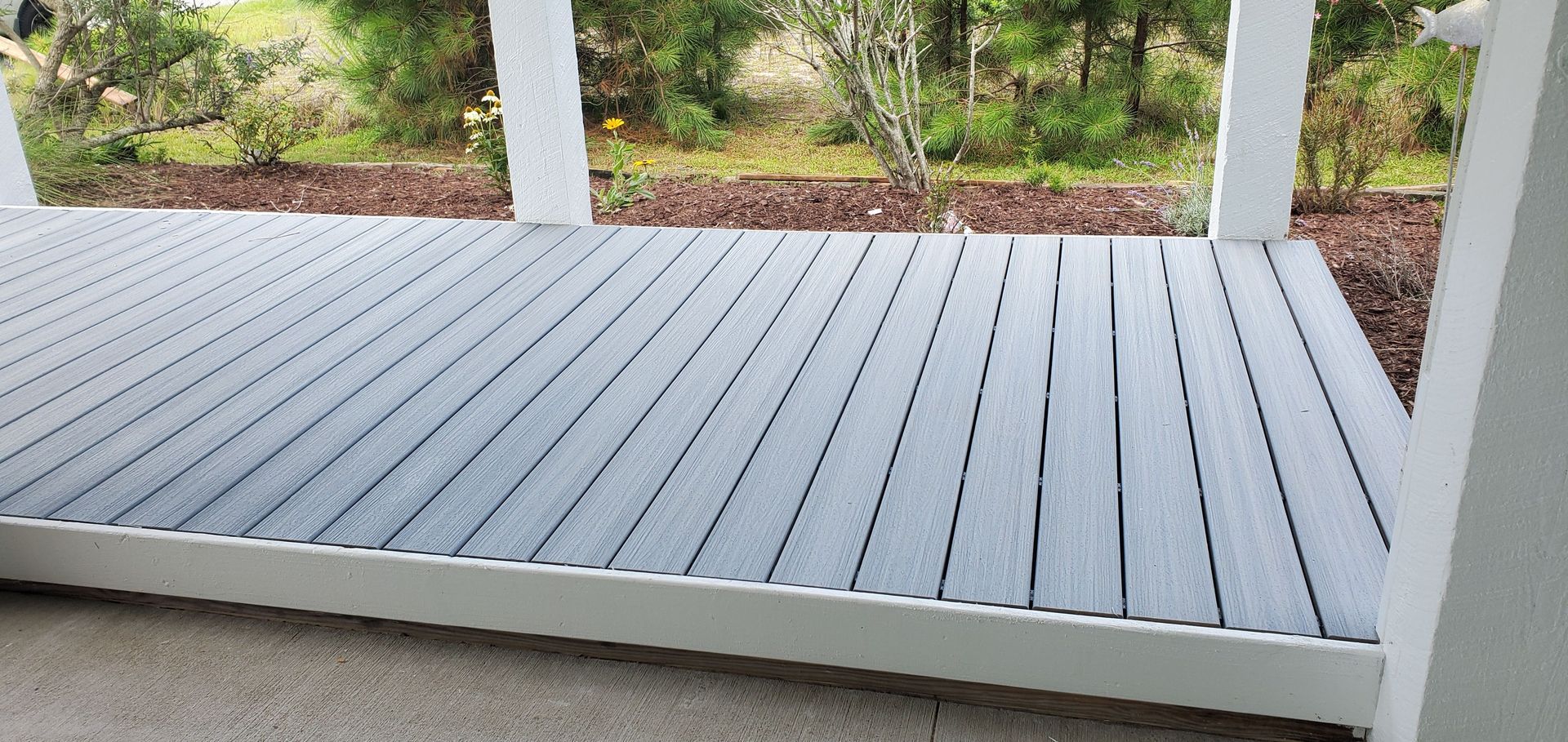 Light gray wooden porch deck with white posts and a view of green shrubs beyond