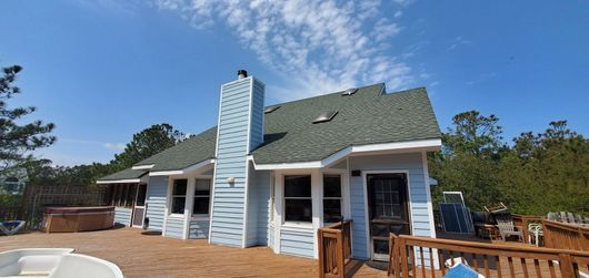 White beach house with green roof and central chimney under a blue sky, with wooden deck and trees nearby