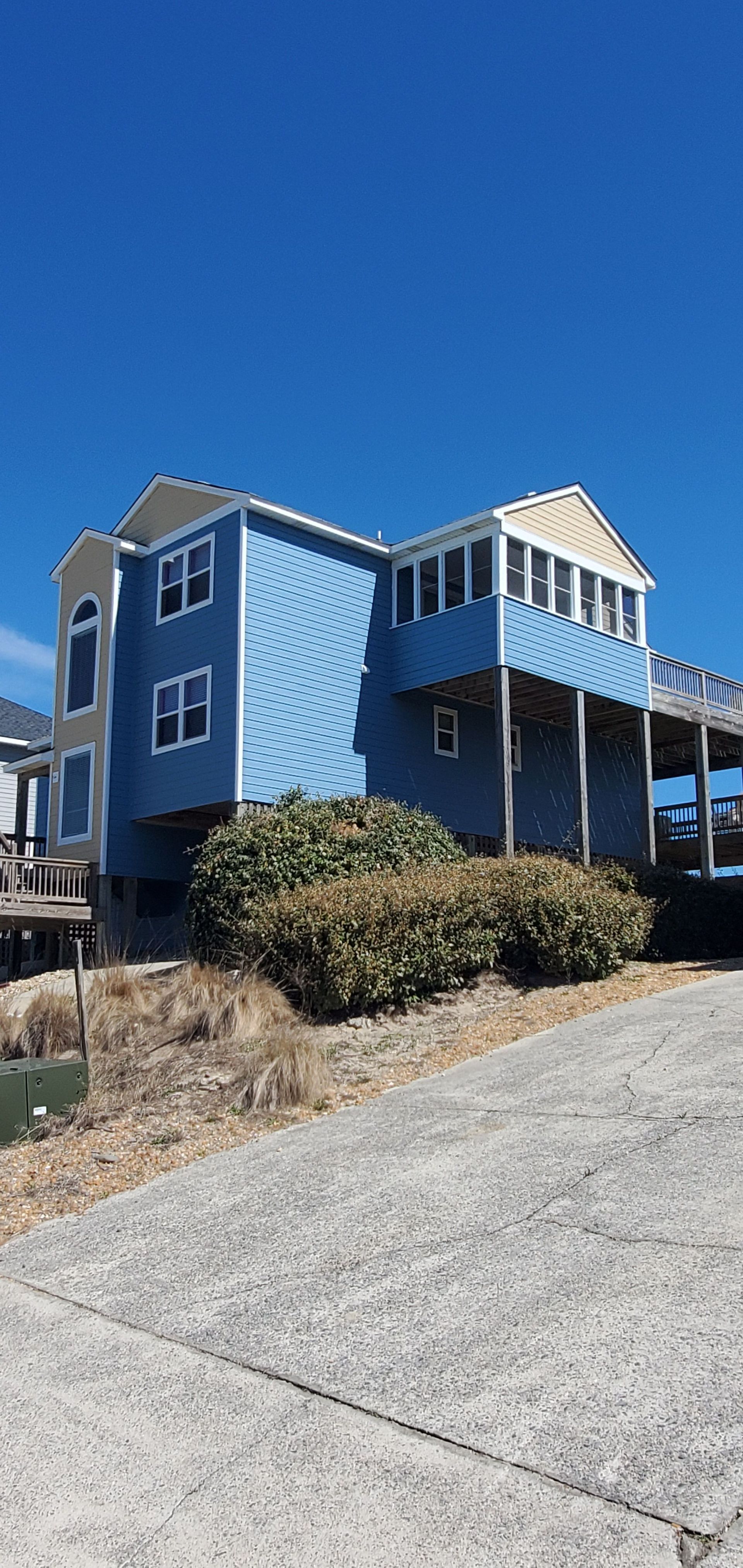Blue house with white trim and a wraparound porch on a sloped driveway under a clear sky