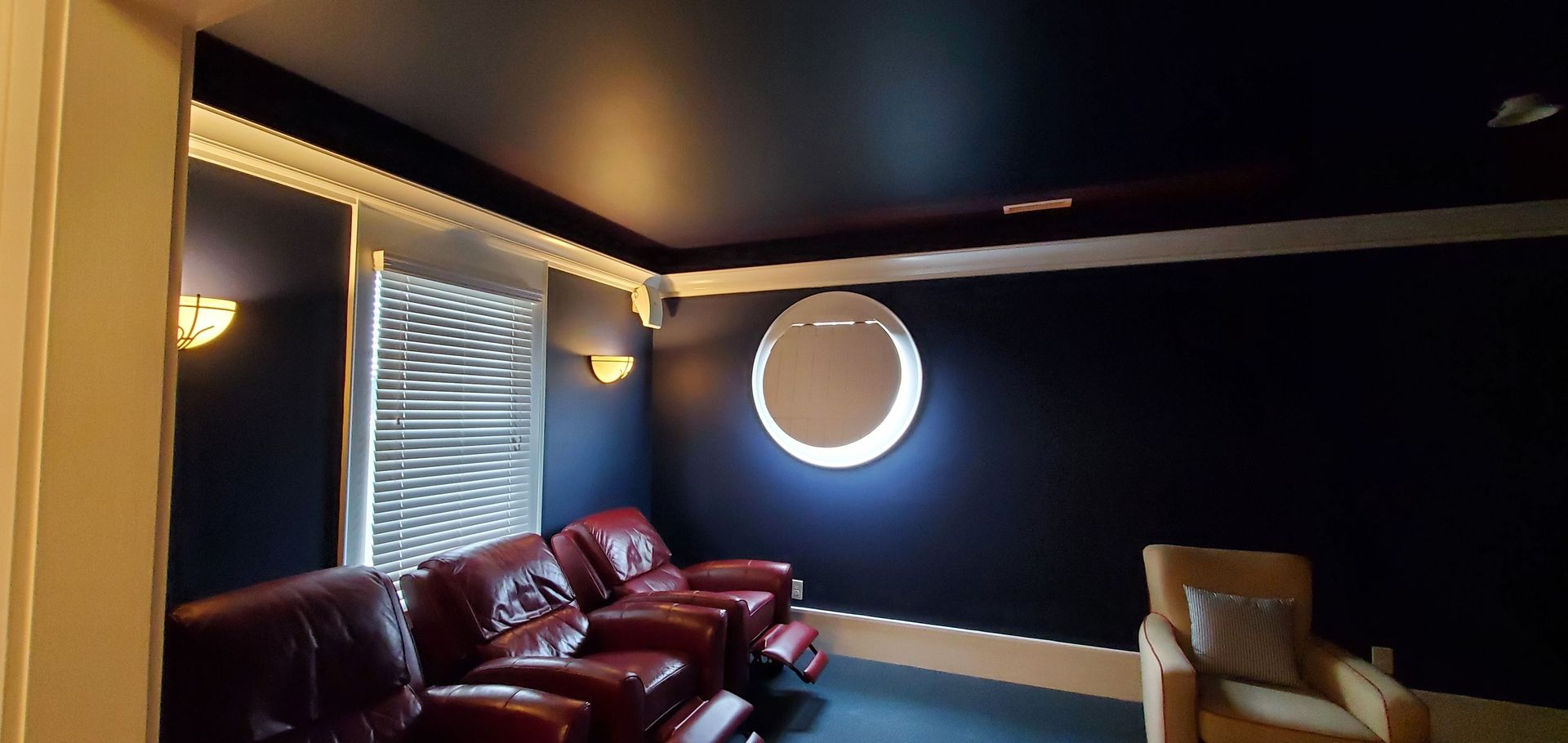 Dim lounge with dark blue walls, red leather sofa, circular wall light, and a tan chair near a window.